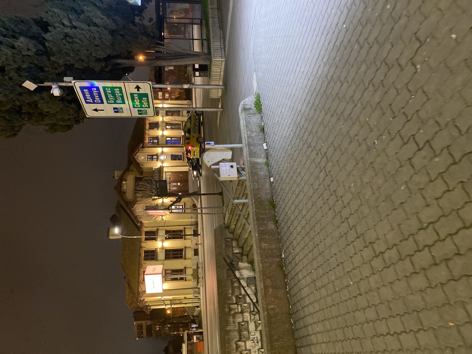 The image shows a street scene at night in Varna, Bulgaria. A multi-story building with illuminated windows and commercial signage is visible on the left side of the frame. Streetlights illuminate the building and street. A road sign indicating directions to "Devnya," "Burgas," and "Sofia" stands in front of the building. The sidewalk on the right side of the frame is paved with interlocking pavers. The scene suggests an urban environment at night. No people are visible in the image.