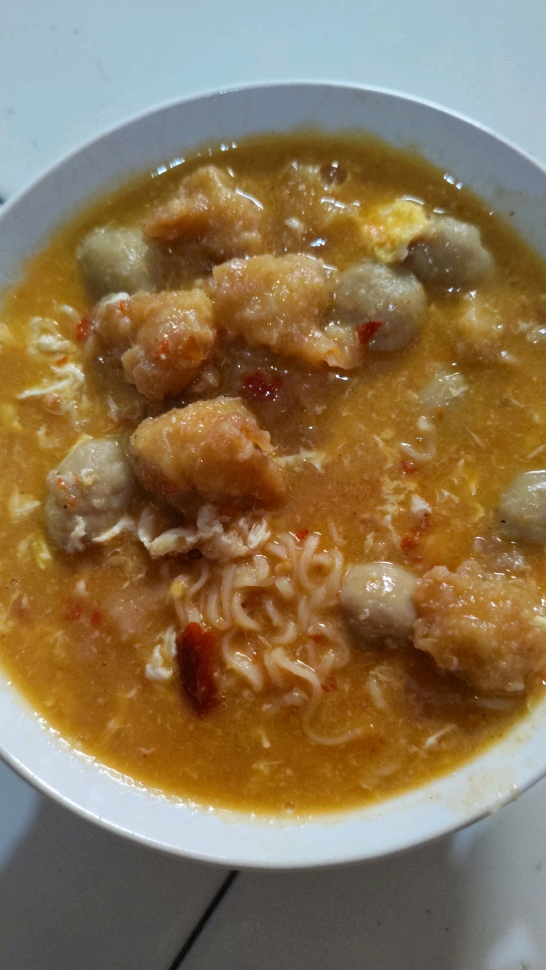 The image shows a close-up, overhead view of a bowl of food, likely a noodle soup dish. The bowl is white and filled with an orange broth. Several pieces of what appear to be fried tofu or chicken are visible, along with round, light-colored meatballs. Thin, wavy noodles are submerged in the broth, and there are also visible shreds of cooked egg and small pieces of red chili. The lighting is somewhat dim, suggesting an indoor setting. The image does not contain any identifiable landmarks or elements that would allow for specific localization within Cimahi, Indonesia.