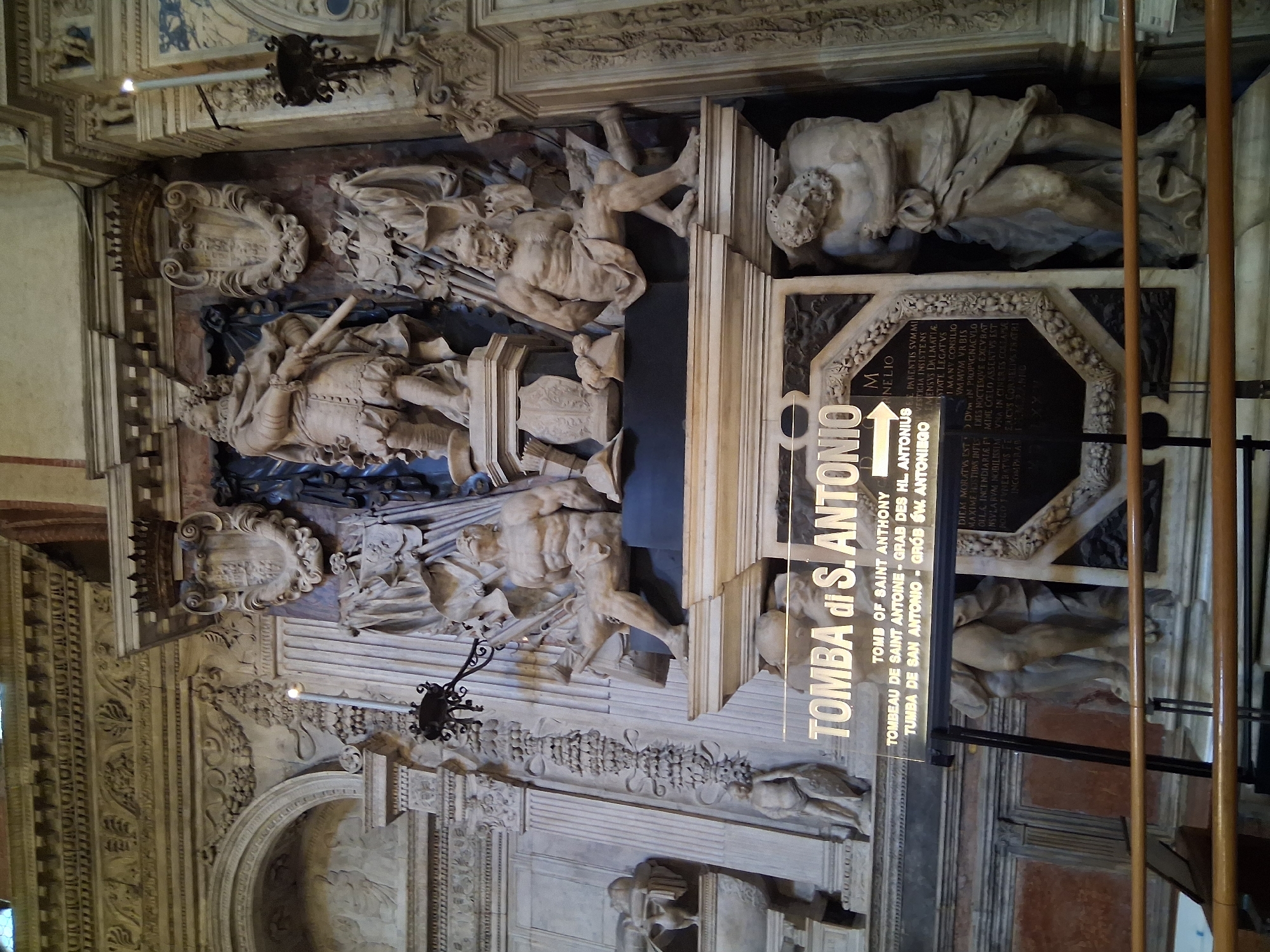 The image shows the tomb of Saint Anthony of Padua, located in the Basilica of Saint Anthony in Padua, Italy. The tomb is a large, elaborate monument adorned with sculptures. At the center of the monument is a statue of a man in armor, holding a sword. Flanking this central figure are statues of muscular men, some seated and some standing. Below the main sculptural group, a sign reads "TOMBA di S. ANTONIO" with translations in several languages. The monument is set against a backdrop of ornate architectural detailing, including a coat of arms and a crown. There are also candles and lamps visible, suggesting this is an interior religious setting. The overall impression is one of grandeur and religious significance.