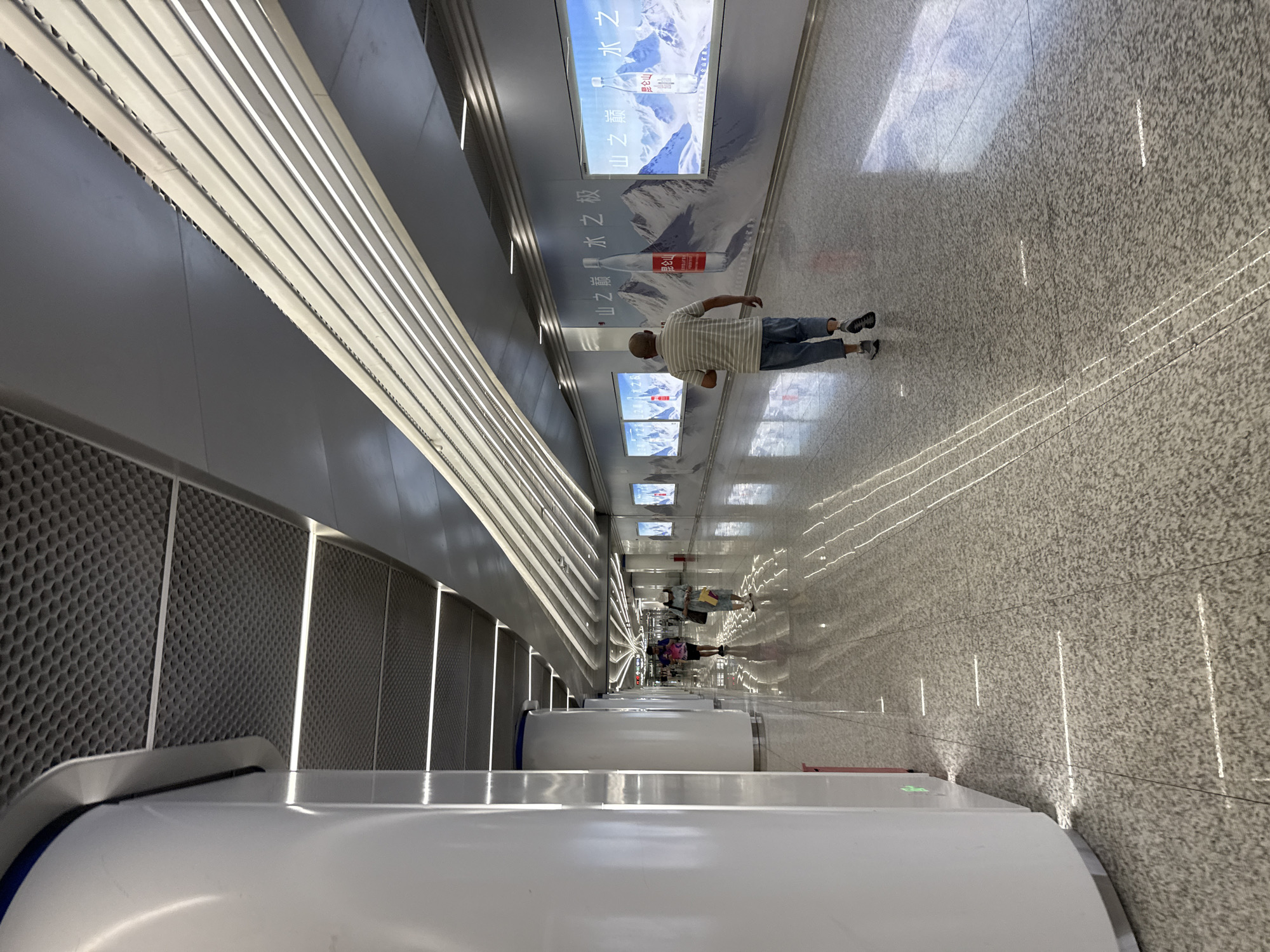 The image depicts an interior view of a modern, well-lit walkway, likely within a public transportation hub or a large commercial building in Haizhu District, China. The architecture features a sleek, minimalist design with a high ceiling. The ceiling is a combination of textured dark gray panels and a series of long, linear light fixtures that cast bright, cool light downwards. The walls are adorned with large, panoramic advertisements featuring mountainous landscapes and bottles of water, which reflect the overhead lighting onto the polished, speckled floor.
In the foreground, a man wearing a horizontally striped t-shirt and denim shorts walks away from the camera. His gait suggests he is moving forward. Further down the walkway, two individuals are visible in the middle ground; one person appears to be wearing a light-colored outfit and a hat, while the other is carrying a bag. Several other figures are present, walking in the same direction or towards the camera, their forms somewhat blurred due to the distance and motion. The floor itself is highly reflective, mirroring the lights and the surrounding architecture, creating a sense of depth and spaciousness. The overall impression is one of cleanliness, order, and contemporary design.