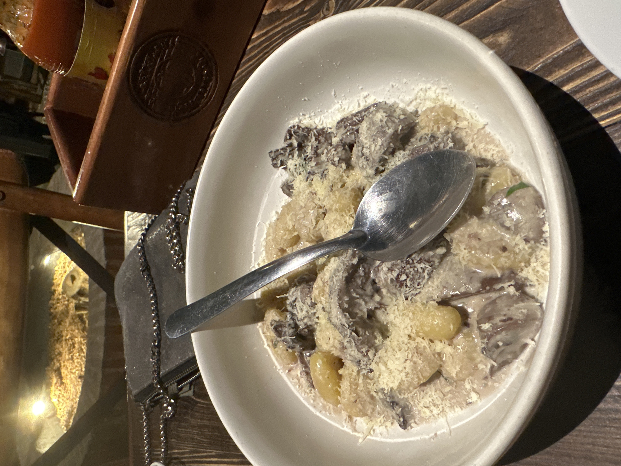 A slightly overhead, angled shot features a white bowl of gnocchi with mushrooms and grated cheese, with a metal spoon resting on top, sitting on a dark wood table. The bowl is centered. In the top left corner, a wooden rack with decorative carvings holds an indiscernible container. To the left of the bowl, a dark-colored purse with a chain strap rests on the table. The setting appears to be a restaurant or dining establishment. The location is the city of San Andrés Cholula, Mexico.