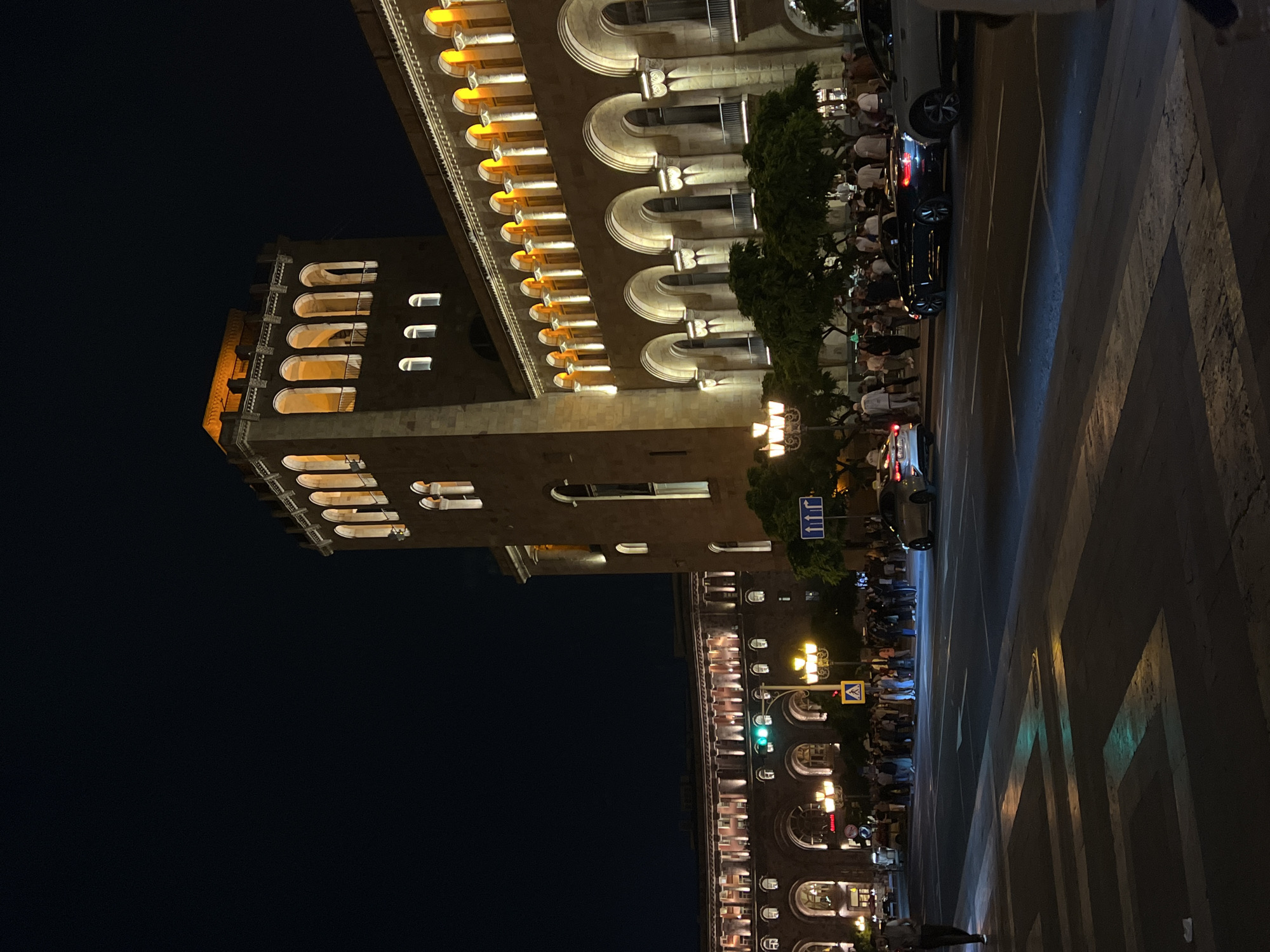 The image shows a nighttime view of Republic Square in Yerevan, Armenia. The illuminated facade of a building with multiple arched windows and details is visible on the right side. People are gathered along the street in front of the building. There are cars on the road. On the left, another building is featured with illuminated arched windows. A traffic light is visible. The street features geometric markings. The sky is dark.