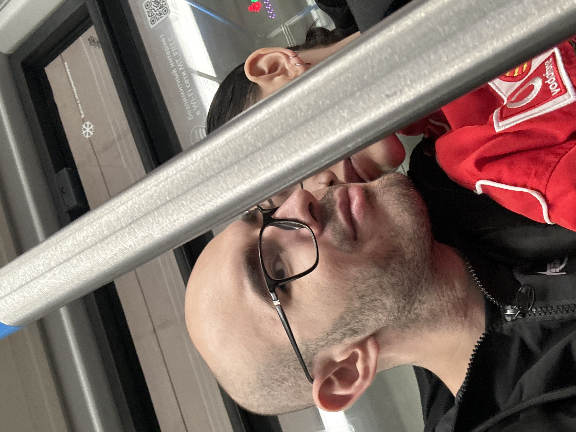 A close-up captures two individuals on public transport. The person in the foreground, likely a male, is wearing glasses and has a visible beard. Behind him and slightly to the side is a person wearing a red jacket with a "Vodafone" patch, partially obscured. The pair appears to be standing or sitting close, possibly conversing. A horizontal metal bar runs across the frame, suggesting it is a grab rail for standing passengers. The background reveals a window with text and a QR code in Russian, indicating the location is likely Moscow, Russia. The text translates to "Free Wi-Fi MT FREE." A snowflake decal is also visible on the window.