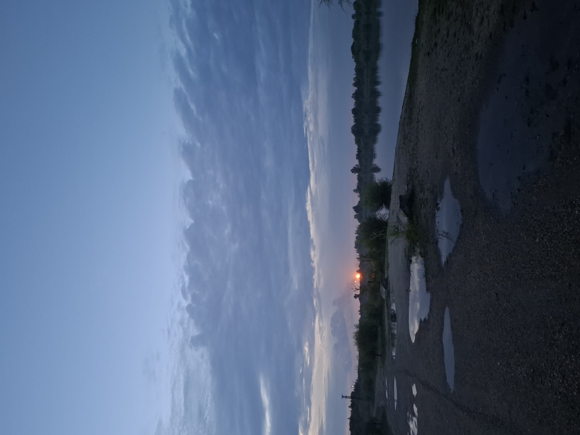 The image depicts a twilight scene at a body of water, possibly a river or lake, with a dirt road in the foreground. The sky is a deep blue with scattered clouds, and the setting or rising sun is visible as a bright orange orb on the horizon, casting a warm glow. The water surface reflects the sky and the distant treeline. The foreground is a rough, unpaved road with several puddles of water, suggesting recent rain. Vegetation, including bushes and trees, lines the opposite bank of the water and forms a silhouette against the sky. A solitary tower-like structure is visible on the left side of the horizon. The overall atmosphere is calm and serene, characteristic of a rural or natural setting at dusk. The scene is consistent with locations along the Amur River in Svobodny, Russia, which is known for its natural landscapes and waterways.