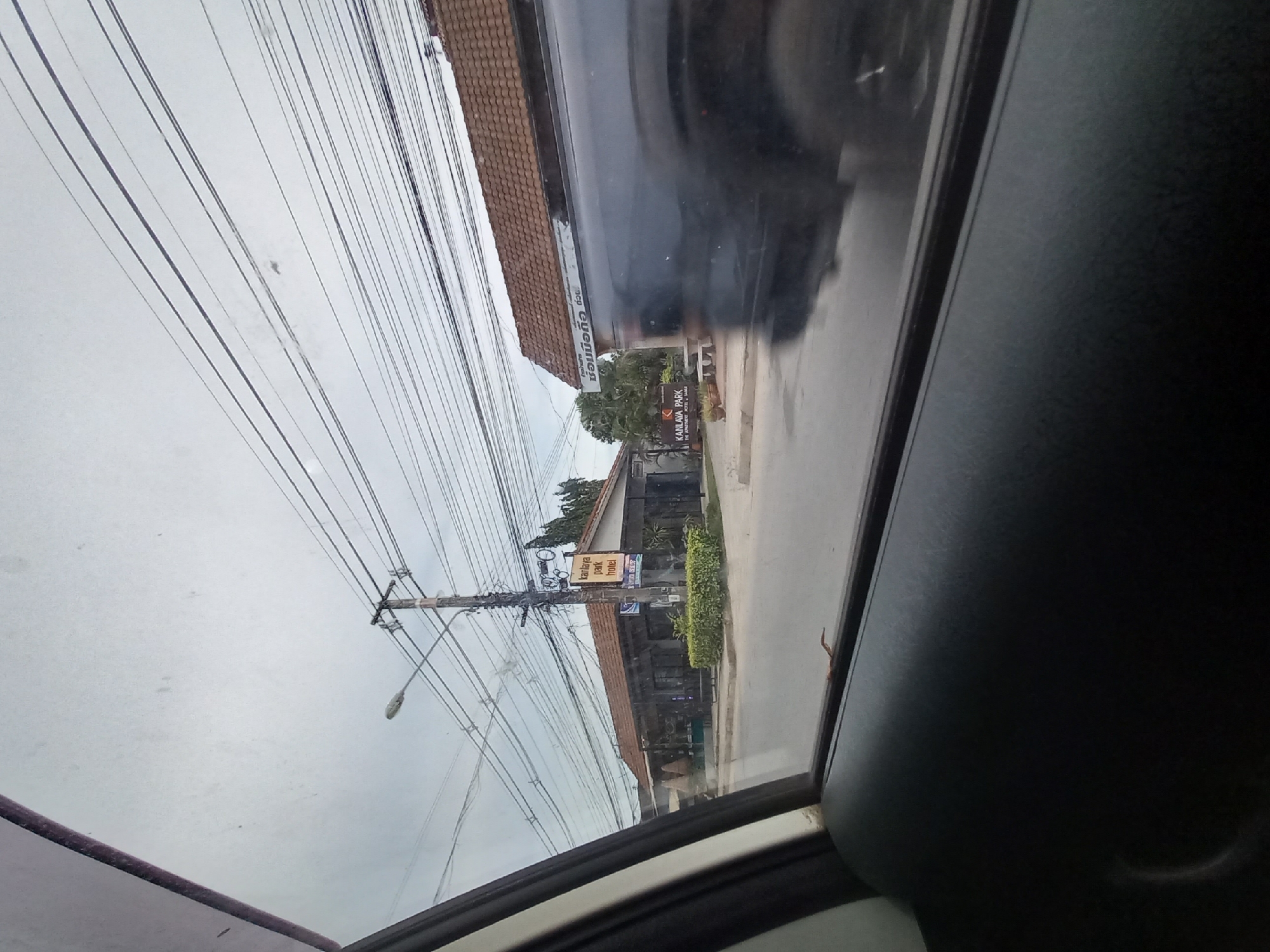 The image is a view from inside a vehicle looking out the side window at a street scene in Baan Tai, Thailand. The sky is overcast. On the right side of the frame, a dark-colored vehicle is partially visible, reflecting the sky and surrounding buildings. In the middle ground, there is a low-rise building with a terracotta tiled roof. A sign on a pole in front of the building reads "kanlaya park hotel" in English. A smaller sign below it has Thai writing. In front of the building, there is a line of green bushes. Further to the left, there is another building, also with a tiled roof. A street light is visible on a pole to the left of the hotel sign. Electrical wires are strung across the upper portion of the image, crossing the sky and obscuring the view of the sky and distant horizon. The foreground of the image shows the interior of the vehicle, with the black leather armrest of the car door visible on the lower left. The overall impression is of a somewhat cluttered urban environment, typical of a street scene in Thailand.