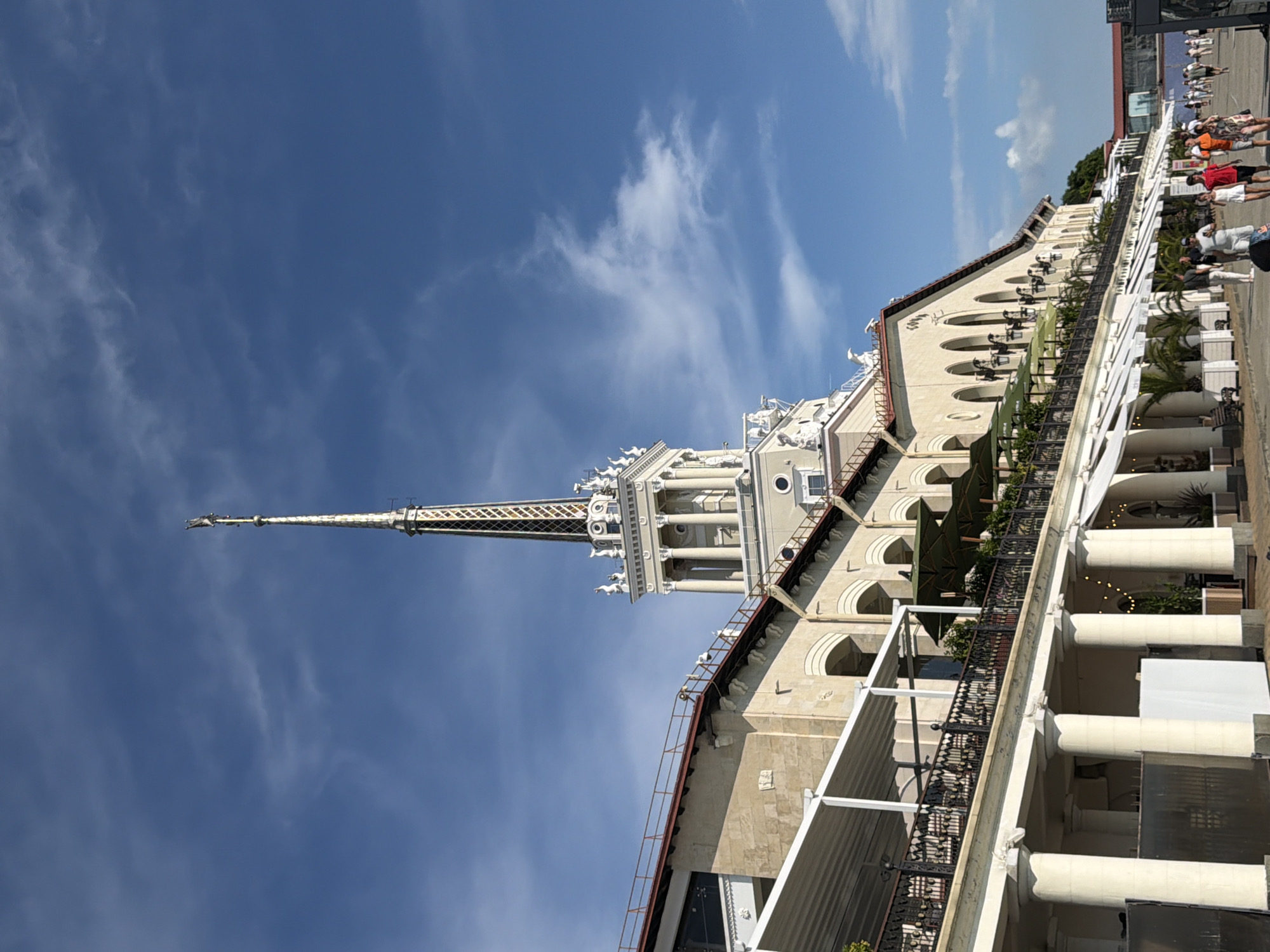 The image depicts a bright, sunny day with a clear blue sky and wispy white clouds. The scene is dominated by a large, ornate building with a tall, slender spire that reaches towards the sky. The building's architecture is classical, featuring arched windows, columns, and decorative elements. Balconies with black wrought-iron railings overlook a walkway lined with more columns and white awnings. Greenery, including palm trees, is visible on the balconies and around the building.
On the walkway, a group of people are casually walking and standing. They are dressed in summer clothing, suggesting a warm day. Their presence indicates that the building is likely a public space, perhaps a hotel, resort, or entertainment venue. The overall atmosphere appears relaxed and leisurely. The image is oriented sideways, with the spire pointing upwards on the left side of the frame. While no specific text is legible, the architecture and the presence of palm trees hint at a warm, possibly coastal, location. Given the context of Sochi, Russia, this could be a building along the Black Sea coast.