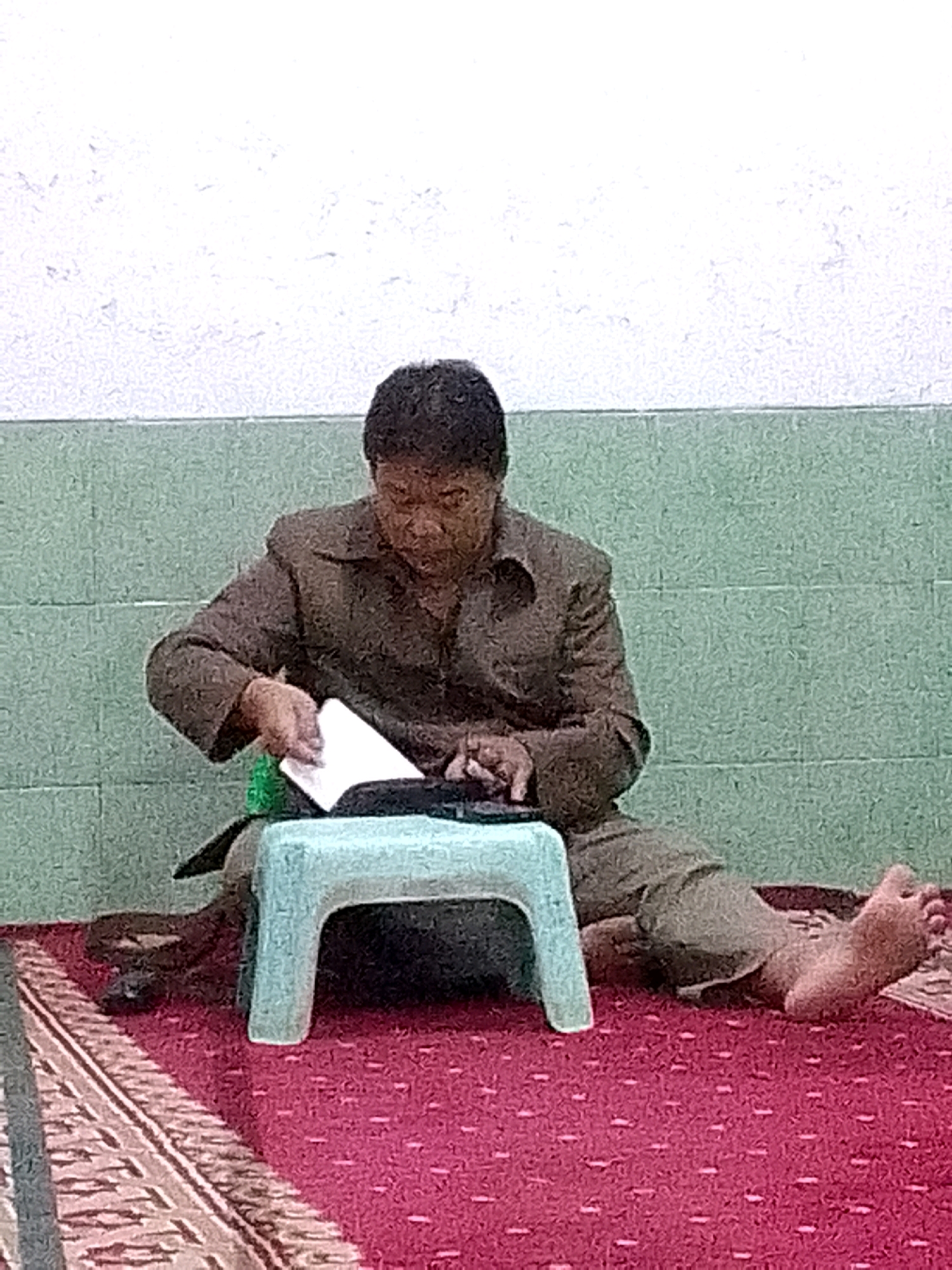 The image depicts a middle-aged man sitting on a red carpet with a patterned border in Gunungmasigit, Indonesia. He is wearing a brown jacket and pants. The man is looking down at a white paper object he is holding in his hands, which rests on a small, light blue stool. His bare feet are visible to his right, with one foot resting on a piece of patterned fabric. The background consists of a textured green wall and a white upper wall.