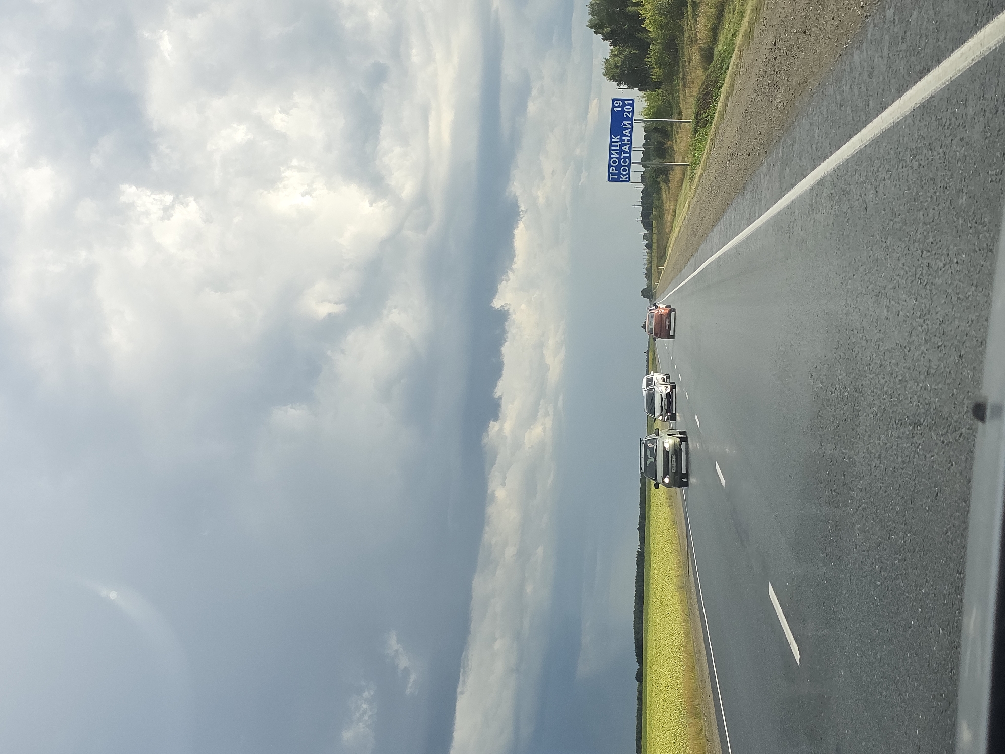 This image depicts a view from a car driving on a wet asphalt road, likely in Russia. The sky is filled with dramatic, dark gray storm clouds, with patches of lighter sky visible. To the left of the road, a vast field of bright yellow sunflowers stretches towards a dark green treeline in the distance. On the right side of the road, a green sign indicates "ТРОИЦК 19" and "КОСТАНАЙ 201" (Troitsk 19, Kostanay 201). Several cars are traveling in the same direction on the road ahead, including a light-colored SUV, a white minivan, and a red sedan. The overall scene suggests a journey through a rural landscape under impending stormy weather. There is no discernible information indicating the location is in the city of Karsy, Russia.