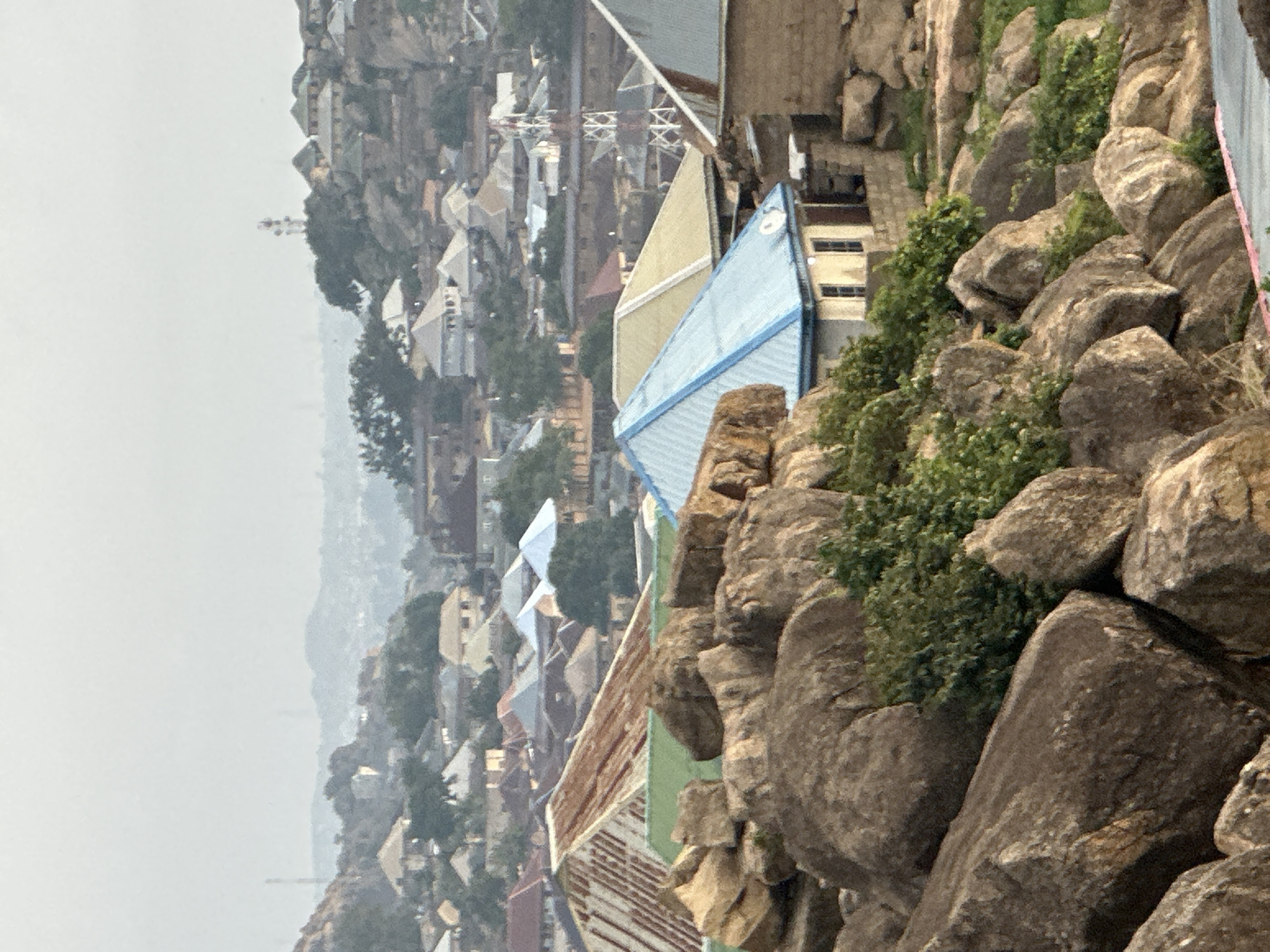 This image, taken in Jos, Nigeria, captures a dense settlement built on and around rocky, hilly terrain under a hazy, overcast sky. The foreground is dominated by large, weathered rock formations and boulders, interspersed with green bushes and small trees.
Beyond the immediate rocky outcrops, numerous residential and commercial structures are visible, extending into the midground and background. Many buildings feature corrugated metal roofs, varying in color from blue, yellow, and light green to rusty brown and unpainted silver. The architecture suggests a mix of older and newer constructions, built closely together, adapting to the undulating landscape. Some structures appear to be multi-story.
In the midground, the urban sprawl continues, with houses densely packed along the slopes. Utility poles or communication towers are discernible among the buildings, indicating infrastructure. The background is characterized by a significant atmospheric haze, blurring the outlines of distant hills and further structures, and creating a soft, diffused light across the scene.
The time of day appears to be daytime, but the heavy haze obscures any direct sunlight or clear shadows, preventing a more precise determination of morning or afternoon. There are no people visible, and no specific activities or events are depicted. No text is visible in the image. The scene conveys the integration of human habitation with a rugged natural environment.
