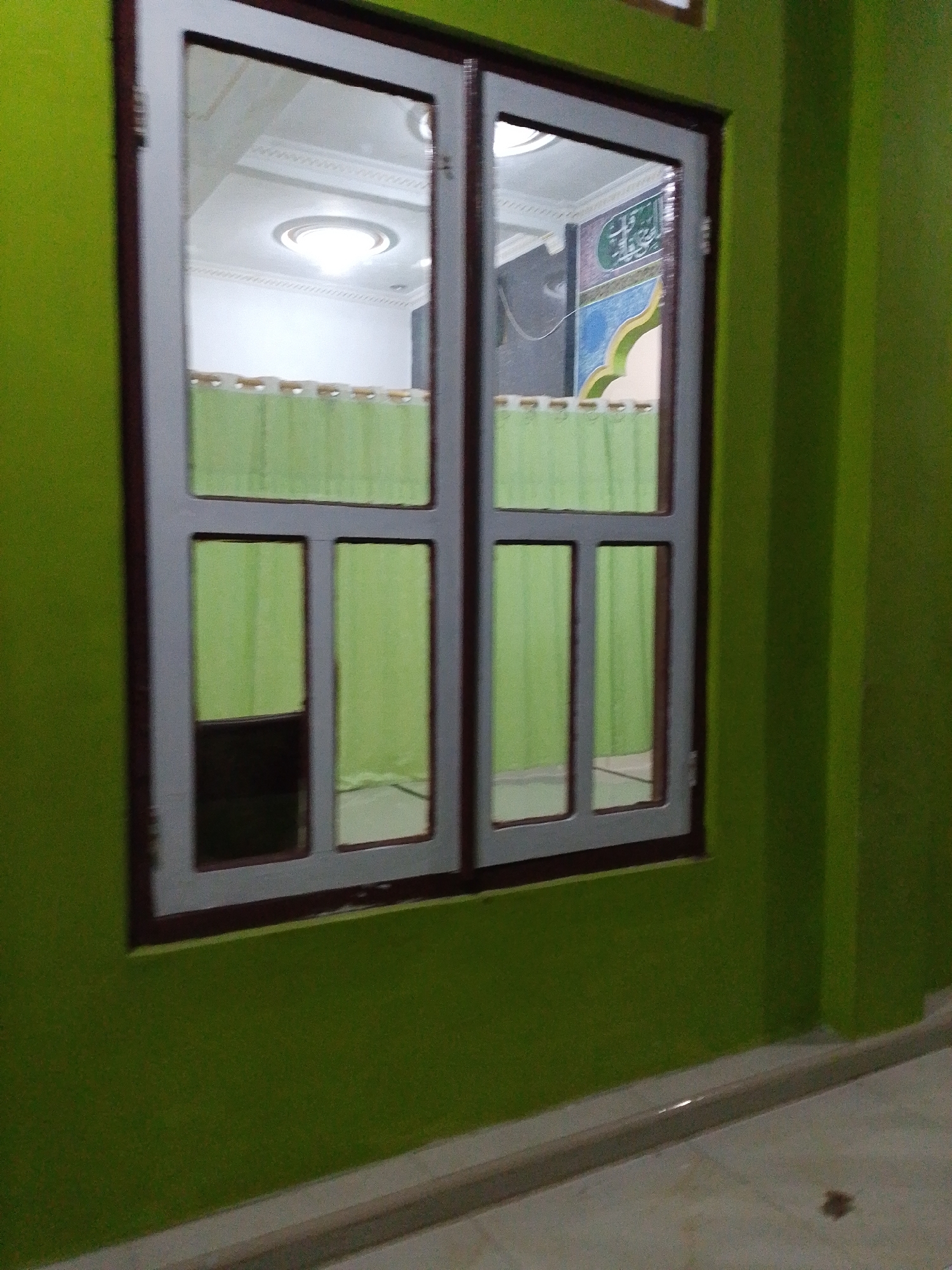 The image shows a close-up view of a window set into a lime green wall. The window has a dark brown frame and is divided into six panes by a white grid. The panes are filled with a light green curtain, except for the bottom left pane which is dark and appears to show the reflection of the room's interior. The window reflects a ceiling with intricate molding and a light fixture. To the right of the window, the lime green wall continues, and a corner of the room is visible. The floor in front of the window appears to be tiled. The lighting suggests the image was taken indoors, possibly in a residential or public building. There are no people or animals visible in the image. The location is identified as Lawatan, Indonesia.
