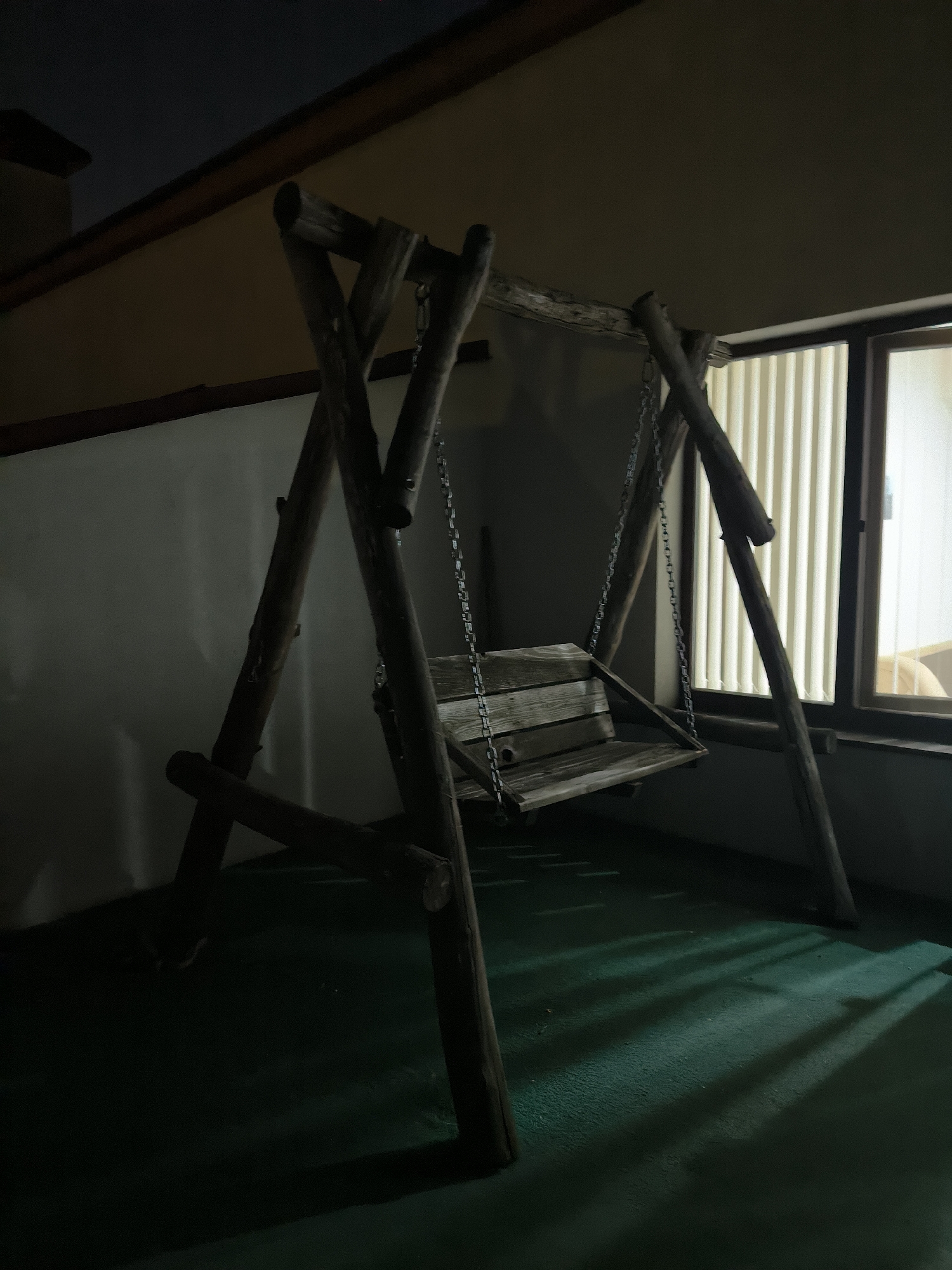 An indoor wooden swing set is pictured. The swing set is made of rough-hewn logs and has a wooden seat suspended by chains. The swing is positioned on a green, textured floor. To the right of the swing, there is a window with vertical blinds, through which light is entering, casting striped shadows on the floor. The background consists of a pale wall and a dark ceiling. The overall lighting suggests it is nighttime or a dimly lit interior. The location is not specified in the image.
