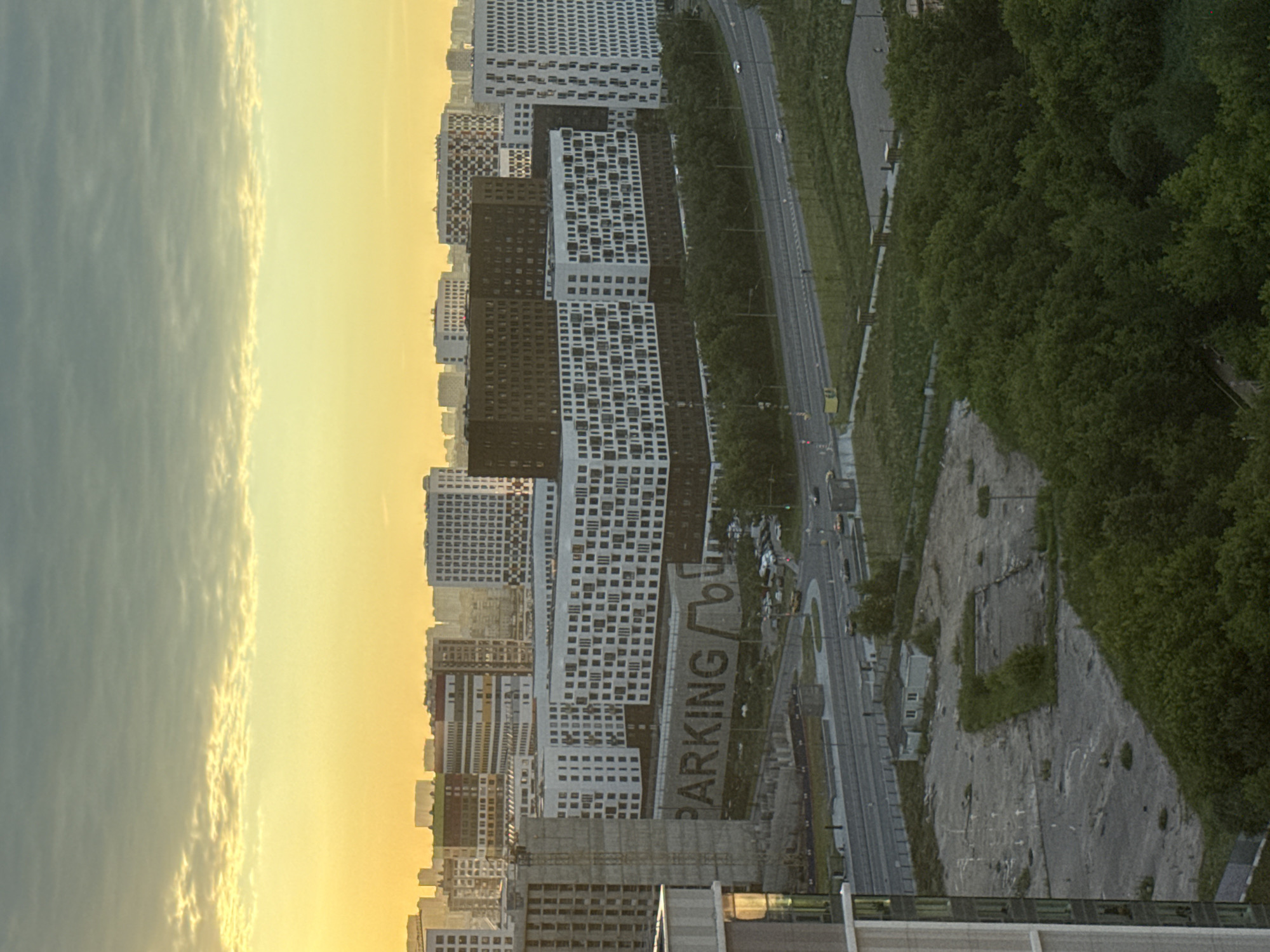 The image depicts a dense urban landscape in Dolgoprudny, Russia, at sunrise or sunset. The sky is filled with a warm yellow and orange hue, indicating either dawn or dusk, with scattered clouds. Dominating the scene are several large, modern residential buildings, characterized by their uniform white and dark facades, featuring numerous rectangular windows.
In the foreground, a partially constructed building or a large concrete structure is visible on the left. Adjacent to this, a long, low-rise building with large, dark letters spelling "PARKING" is prominent. This structure appears to be part of a larger complex. A road runs in front of these buildings, with several vehicles, including cars and a yellow truck, visible.
Beyond the immediate structures, the cityscape extends with more residential blocks and buildings of varying heights and architectural styles. A strip of dense green trees separates the road and parking area from a field of undeveloped land with scattered concrete foundations and a small, low-lying building with a gabled roof. The overall impression is one of a developing residential area within a larger city. No people are clearly discernible in the image.