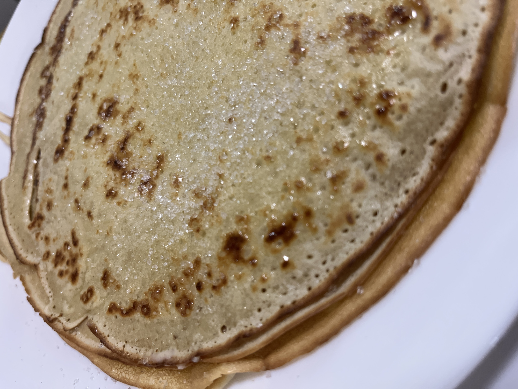 A close-up, overhead shot shows a stack of crepes on a white plate. The crepes are golden brown with darker caramelized spots and sprinkled with sugar crystals, which glint in the light. The edges of the crepes are thin and slightly irregular. A few stray sugar crystals are scattered on the white plate to the left of the crepes. There are no people or animals visible in the image. The lighting appears to be from above, highlighting the texture and shine of the sugar. The image does not contain any visual information that would suggest a specific location in Krasnyye Barrikady, Russia.