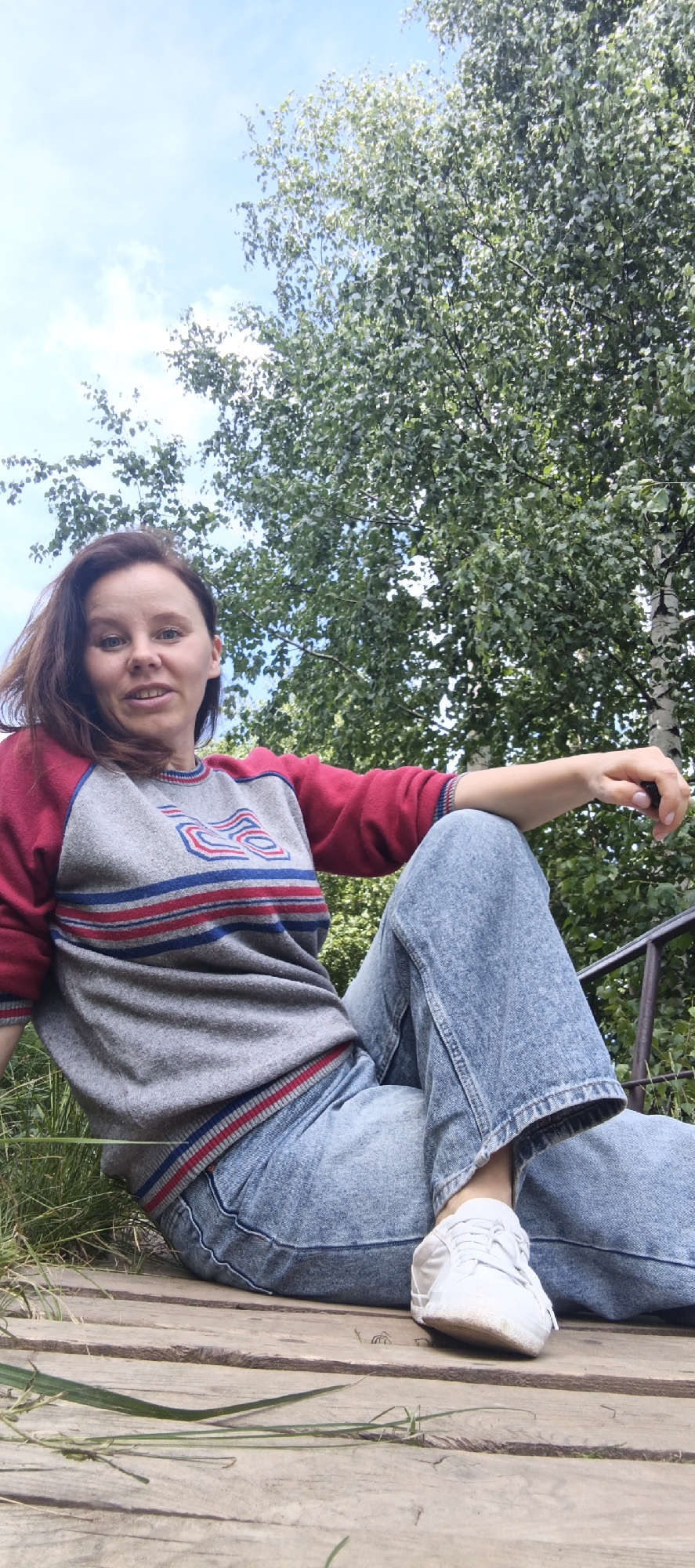 An outdoor, low-angle, full-body shot shows a woman sitting on a wooden platform. She has brown hair and is wearing a gray sweater with red sleeves and blue and red stripes across the chest. She is also wearing light blue jeans and white sneakers. The woman is smiling and looking at the camera. The background consists of green trees and a blue sky with some clouds. The location is Unknown, Unknown.