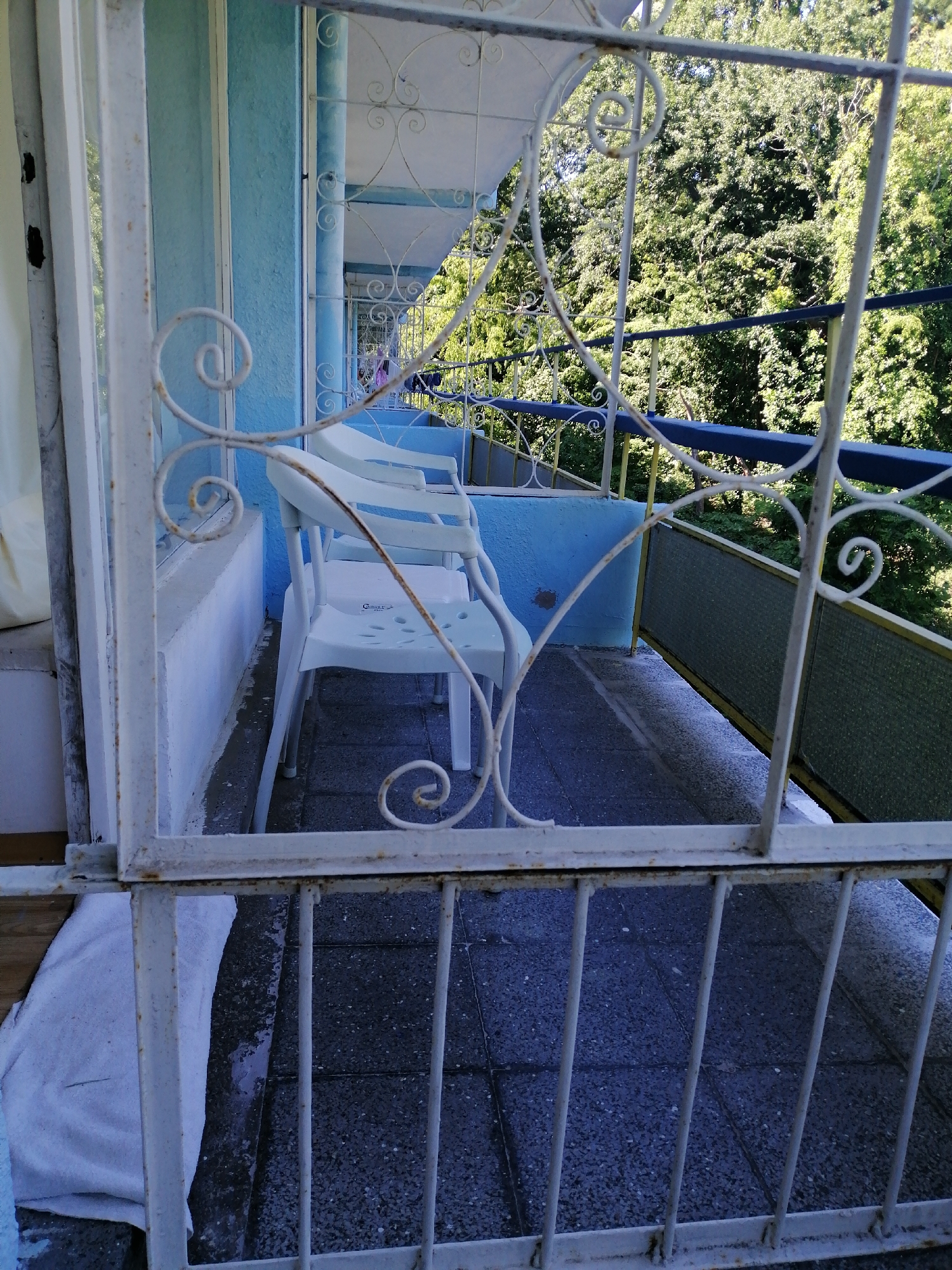 The image depicts a balcony area in Varna, Bulgaria, with a pale blue stucco wall and a tiled floor. White metal railings, some with decorative scrollwork, frame the scene. Two white plastic chairs are visible on the balcony, one placed slightly behind the other. Through the railings and an open doorway, a lush green forest canopy can be seen. The overall impression is of an outdoor space that is slightly weathered, with a hint of domestic use suggested by a white towel folded on the floor near the entrance.