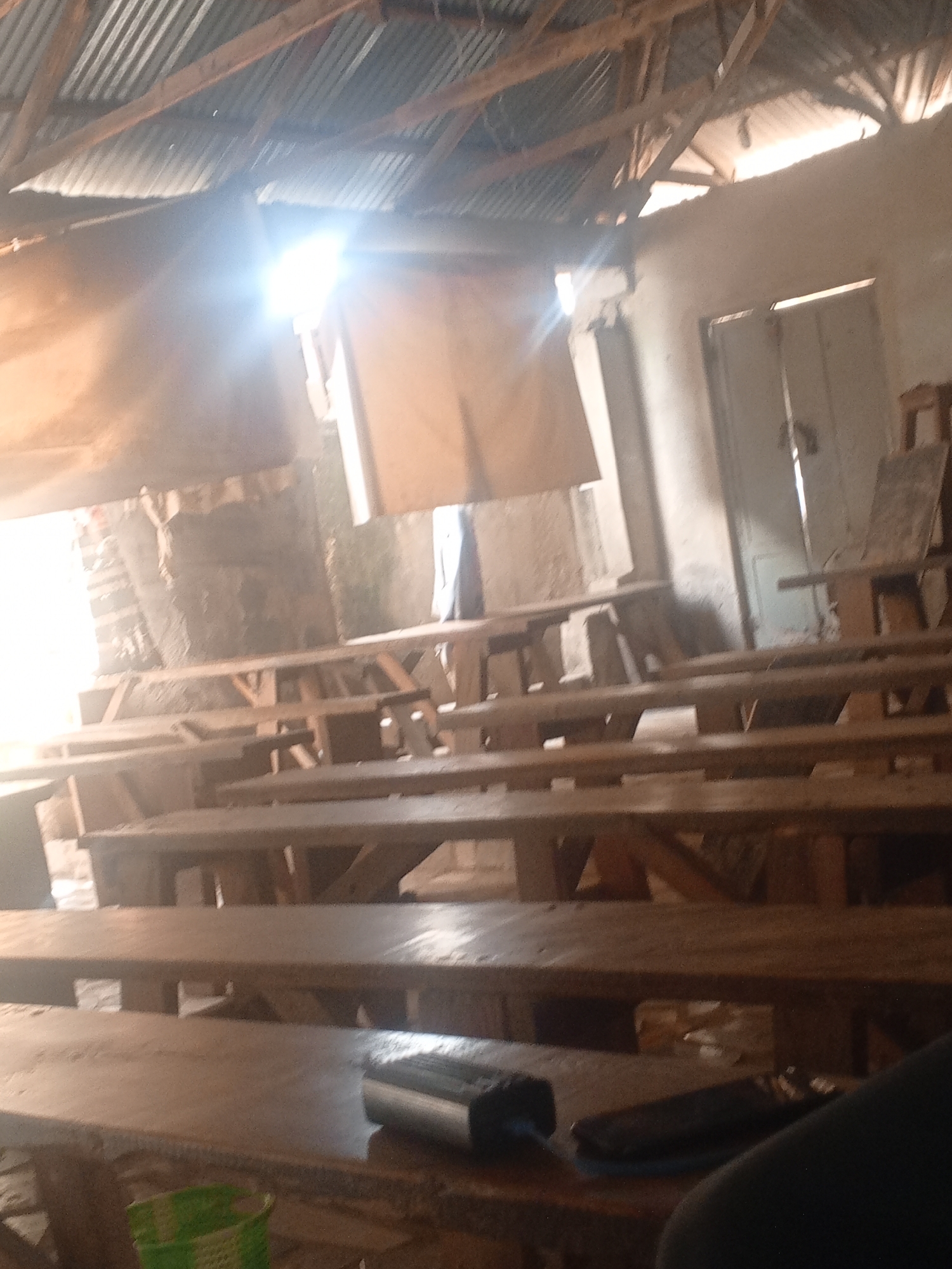 The image depicts the interior of a classroom with a corrugated metal roof and unfinished wooden desks. Sunlight streams in from an opening in the roof, creating bright glare. The room appears to be sparsely furnished with rows of wooden benches and desks. There is a door visible in the background. The overall scene suggests a basic and possibly rural educational setting. The lighting indicates it is daytime, likely morning or afternoon, with bright sunshine. No people are visible in the frame, and there is no discernable text. The condition of the room, with exposed beams and makeshift construction, suggests it may be a temporary or underdeveloped school facility. The context provided places this scene in Jalingo, Nigeria.
