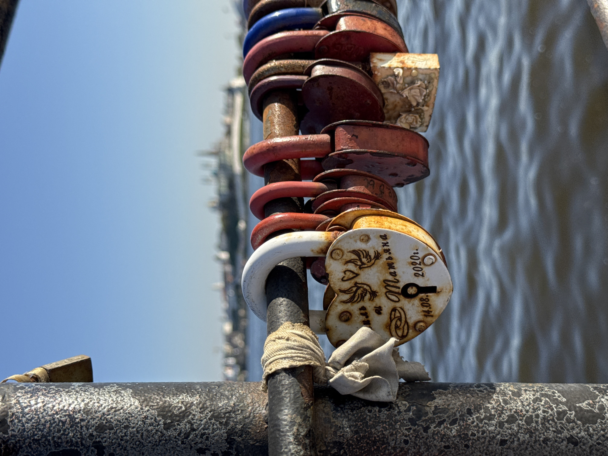 The image shows a close-up of a metal railing at the Millenium Bridge in Kazan, Russia, on which a collection of love locks has been attached. The locks are of various shapes, sizes, and colors, mostly rusted. The focal point is a white heart-shaped lock with the name "Мальвина" (Malvina) and the date "14.08.2020" engraved on it. A piece of cloth is tied around the railing near the lock. In the background, the water of the Kazanka River and the city skyline can be seen under a clear blue sky.