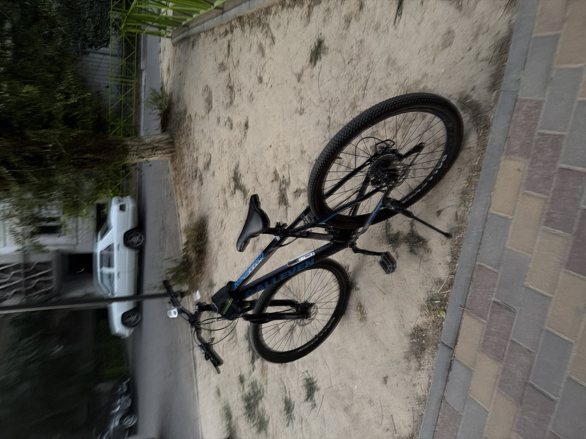 A black mountain bike with blue accents is parked on a sandy surface next to a paved walkway in Shymkent, Kazakhstan. The bike has disc brakes and a kickstand. In the background, a white sedan is parked on a street, and a building with an unusual architectural style is visible. A green fence runs along the side of the street. The overall lighting suggests it is either early morning or late evening.