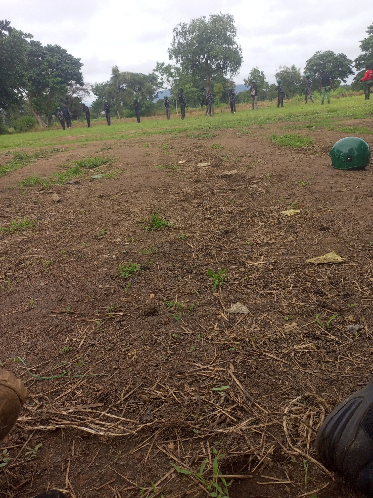 The image depicts an outdoor scene in a grassy field with a dirt path in the foreground. A line of individuals, appearing to be military personnel in uniform and some wearing green helmets, stand in formation on a grassy incline in the background. They are armed with rifles. A green military helmet lies on the dirt path in the foreground, to the right of the frame. The environment consists of trees and vegetation, with a cloudy sky overhead. The scene is likely a training or operational area within or near the city of Takum, Nigeria.