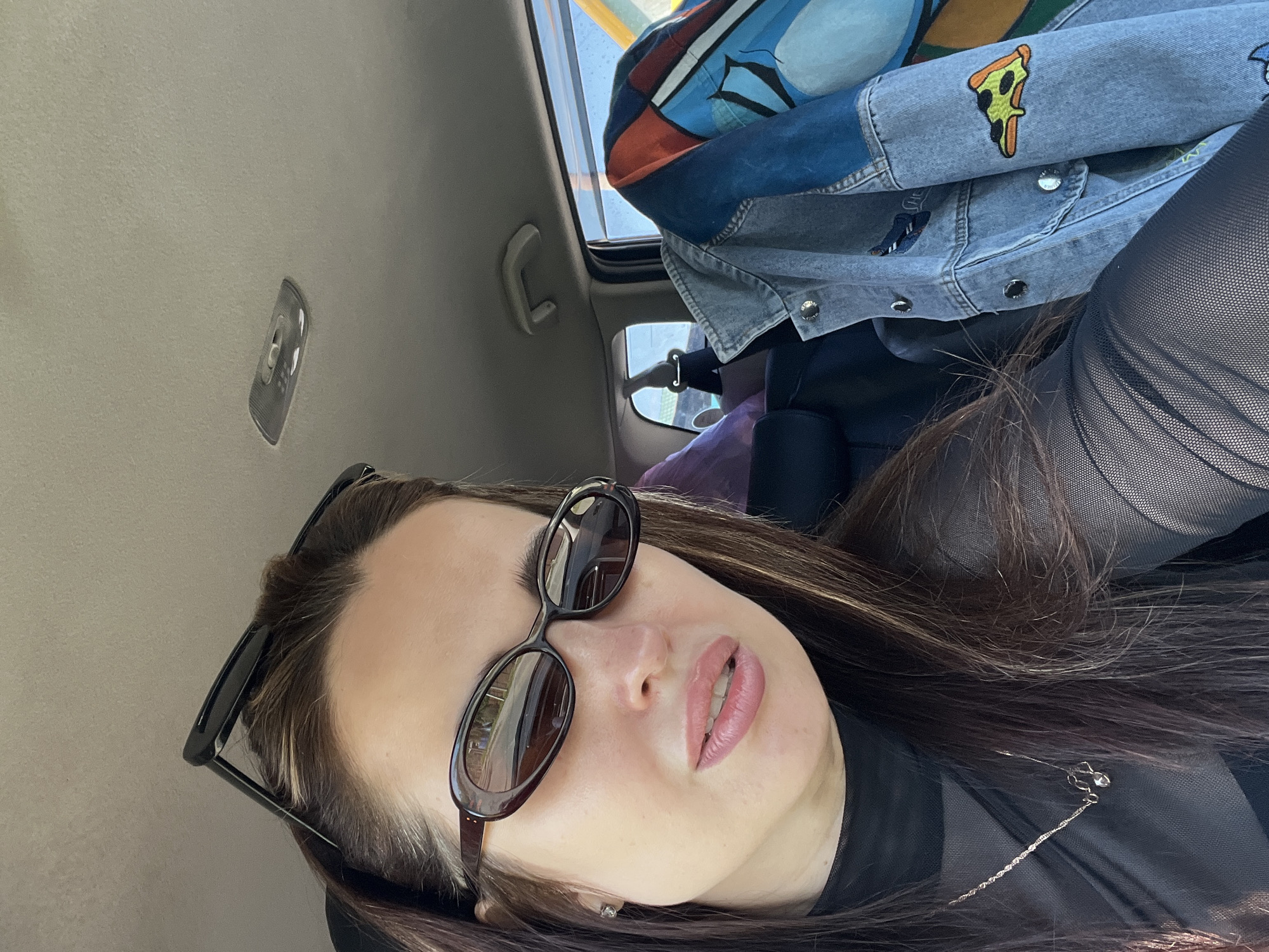 A selfie taken from inside a car. A woman with long brown hair, wearing sunglasses and a black top with a silver necklace, is looking up at the camera. Above her is a jacket with colorful patches and a pizza slice design. The car has a sunroof and a gray interior. The location is the city of Unknown, Unknown.