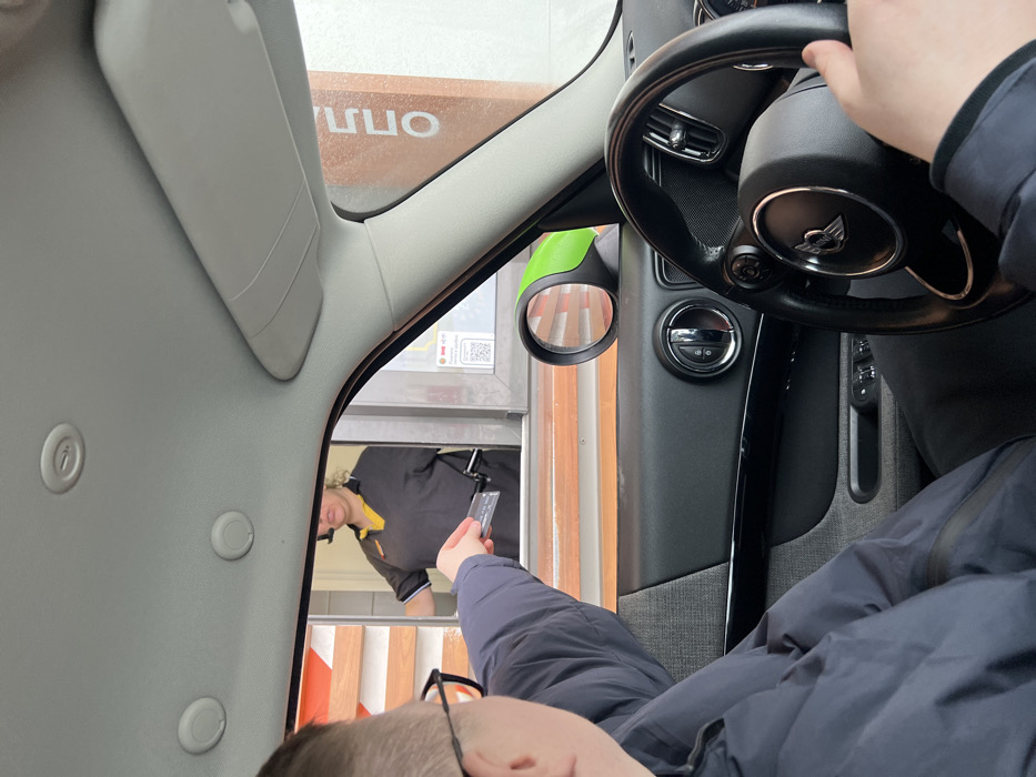 The image depicts a transaction occurring at a drive-through service window in Elino, Russia. A person, seated in the driver's seat of a Mini Cooper, extends their right arm through the open window, holding a dark rectangular payment card. The individual is wearing a dark blue jacket and glasses, with the back of their head and shoulder visible in the foreground. Their left hand grips the steering wheel. Through the vehicle's window, a female employee is visible, wearing a dark grey polo shirt with yellow accents and a headset with a microphone. She stands inside the drive-through facility, appearing to reach for or accept the payment card. A transparent payment tray or device extends from her side. The exterior of the drive-through facility features an orange-toned window frame and a vertical orange panel with partially visible white Cyrillic letters "ОППЛ". A white sign below the window displays a QR code and Russian text, including "Сбербанк Pay" and "Оплата по QR-коду," along with a "Jeep SberPay" logo, indicating electronic payment options. The vehicle's interior shows parts of the dashboard, door panel, and headliner, consistent with a modern car.