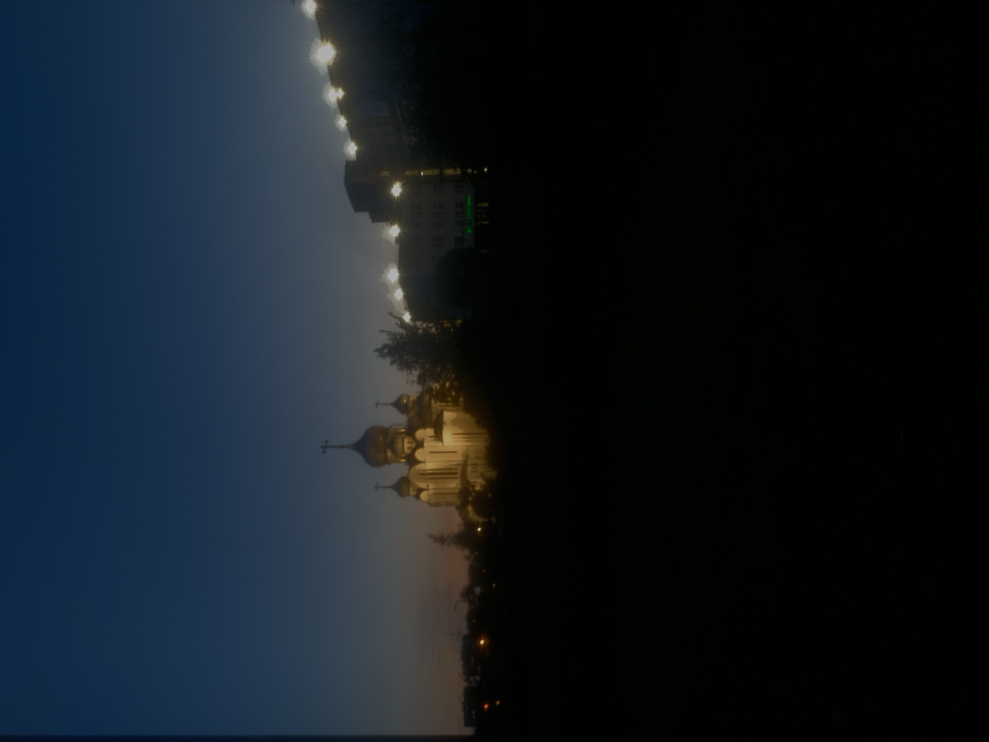 This image, captured at dusk in Vsevolozhsk, Russia, shows a dimly lit scene featuring a prominent church with golden domes and a modern building with bright lights. The church, a traditional Orthodox structure, is illuminated, casting a warm glow against the darkening sky, and is surrounded by trees and foliage. To the right, a multi-story building exhibits a series of bright, star-like lights along its roofline, and smaller illuminated windows are visible on its facade. The foreground is entirely consumed by darkness, obscuring any ground-level details. The overall impression is of an urban landscape at twilight, with religious and secular architecture juxtaposed against a deep blue and orange gradient sky.