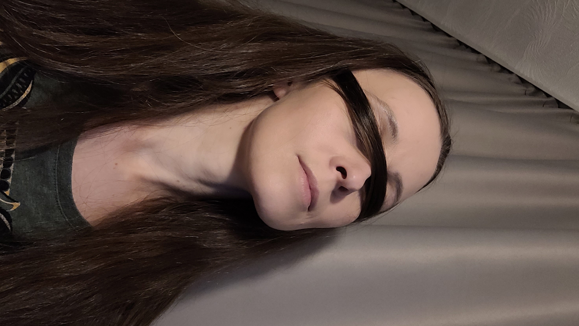 A close-up, chest-up, medium shot shows a fair-skinned woman from the chest up, her head tilted slightly upwards and to the right. Her long, dark brown hair is pulled forward, covering her eyes and forming a dark band across her face. Her lips are slightly parted, and her eyes are not visible. She wears a dark green t-shirt with a patterned neckline. The background consists of soft gray curtains that are drawn, with a faint texture visible. The lighting is dim, casting soft shadows on her face and neck. The image appears to be taken indoors.