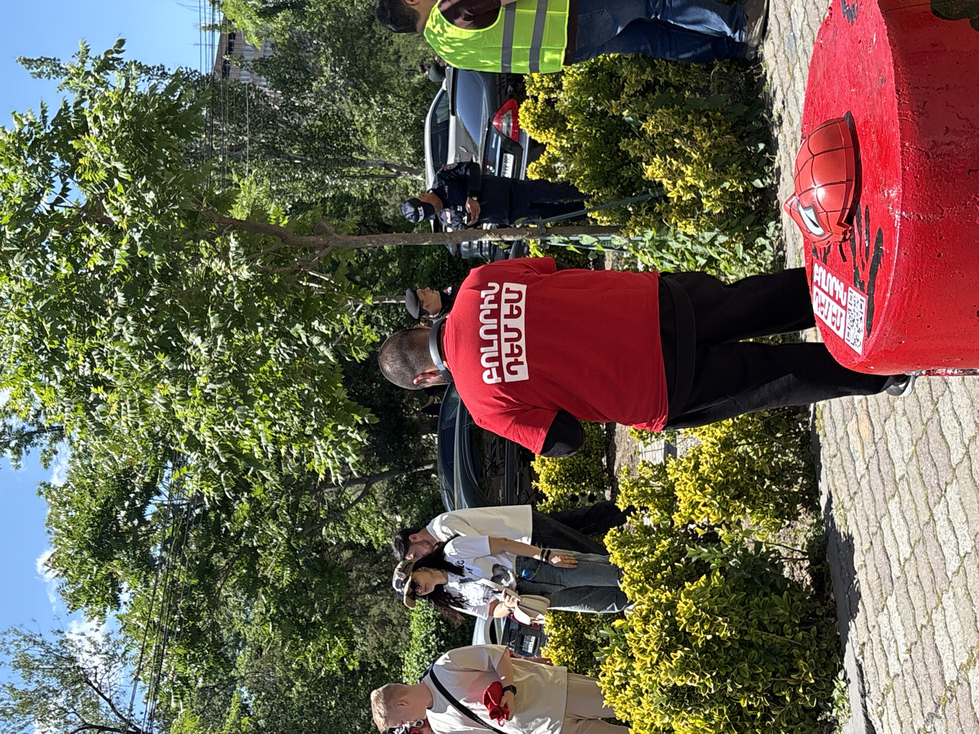 The image captures an outdoor scene on a paved area in Yerevan, Armenia. A red cylindrical object with a Spider-Man mask affixed to it stands on the right. A man wearing a red T-shirt with white lettering in Armenian stands next to it. In the background, there are several people, including one wearing a reflective vest, a person in a police uniform, and others. Trees and green bushes are dispersed around the scene. Several parked cars are visible in the background. Overhead power lines stretch above the trees.