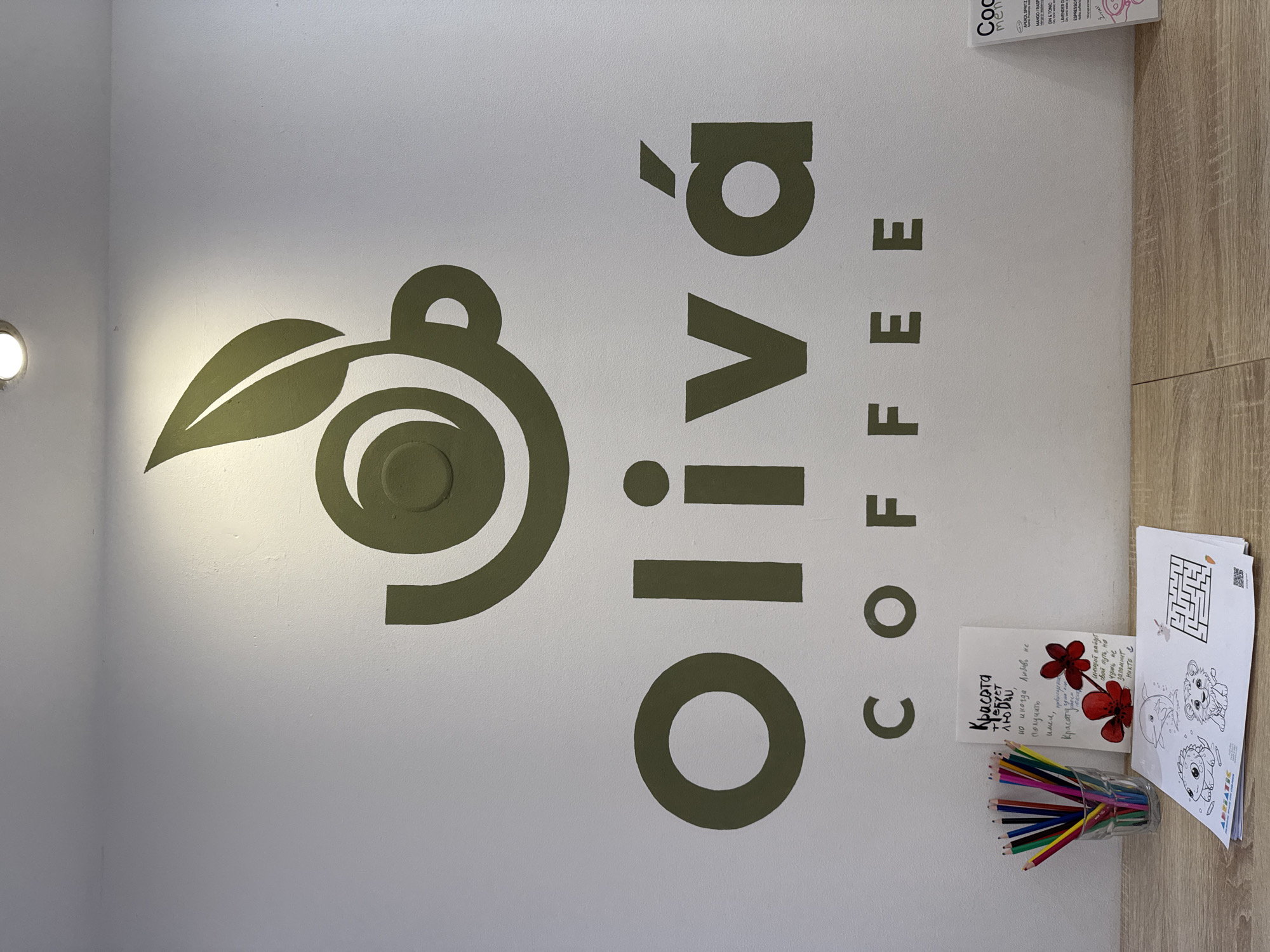 The image shows a wall with the logo of "Olivá Coffee" vertically written in olive green color. The logo includes a stylized olive branch on the left. The photograph is taken inside a building, likely a coffee shop or cafe. There are coloring pencils in a glass, along with printed coloring sheets and a card with text in Russian, placed on a wooden surface next to the wall. The location of the image is likely in Budva, Montenegro, given the mention of "Živjeli" on the printed menu card.
