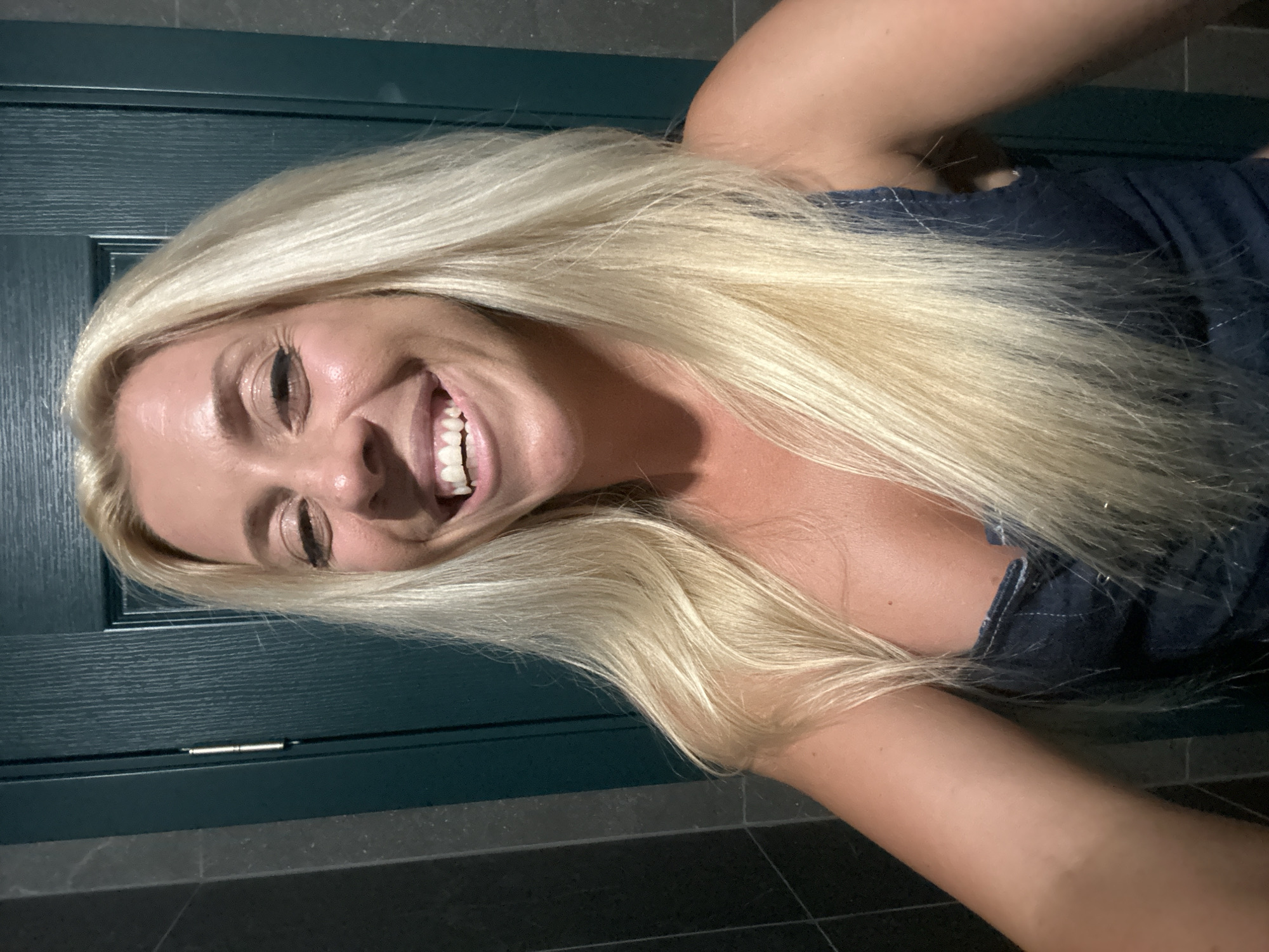 A blonde woman is taking a selfie in what appears to be a bathroom. She is smiling broadly, with her mouth open and teeth visible. She is wearing a dark blue denim top. The background includes a dark green door and dark tiled walls. The overall lighting suggests an indoor setting, possibly at night. There is no information in the image that would indicate the location is Pazardzhik, Bulgaria.
