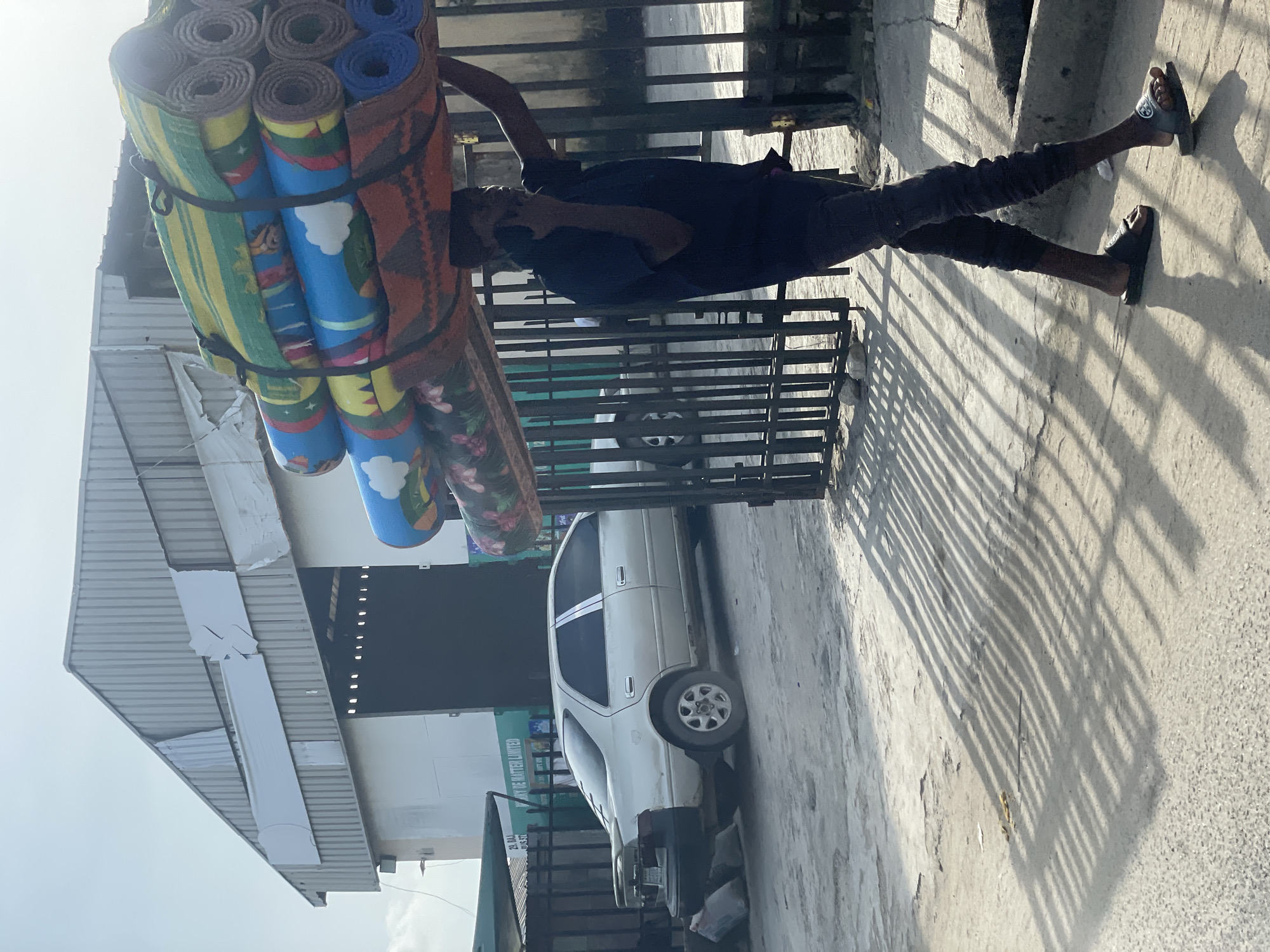 A man in Ado, Nigeria, carries a stack of rolled mats on his head while standing on a concrete sidewalk. He wears a dark blue shirt and dark pants, with sandals on his feet. The mats are secured with black straps and feature various colors and patterns, including depictions of cartoon characters and floral designs. The location shows a street with a white car parked next to a building with a corrugated metal roof and advertising signage. A metal gate casts shadows on the sidewalk.