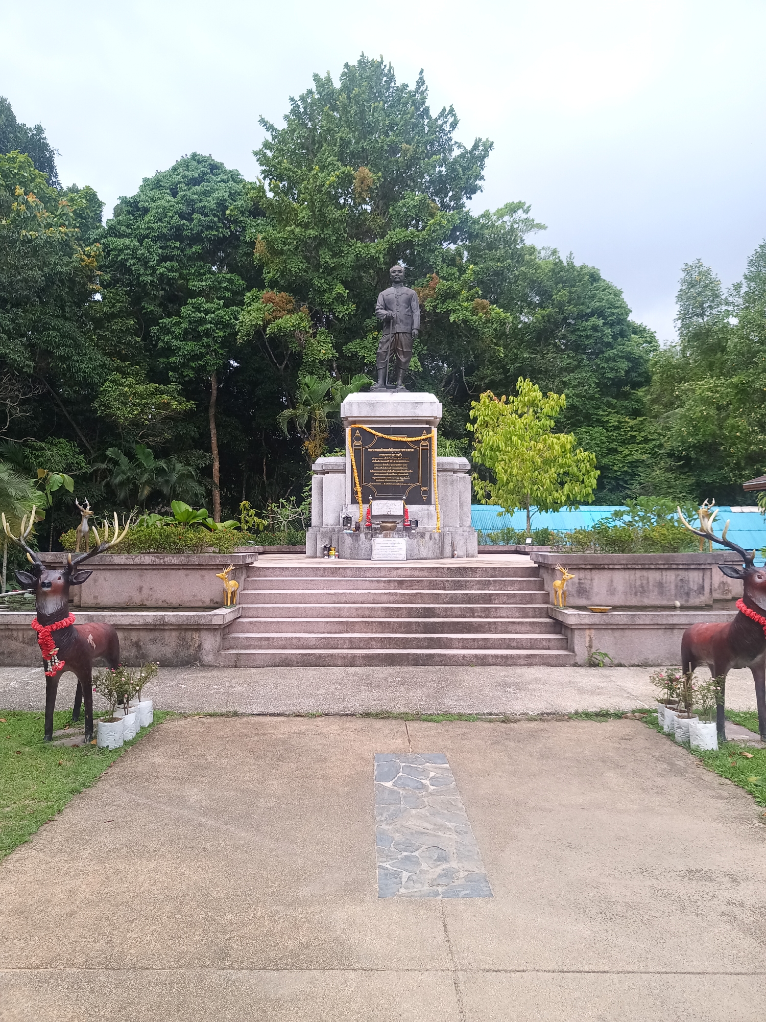 The image shows a statue of a man on a pedestal in Baan Thong Nang, Thailand. The statue stands atop a series of steps leading up to a concrete structure. The pedestal features a plaque with text in Thai. In front of the steps are two deer statues, each adorned with a red floral garland. The ground is paved, with greenery visible to the sides. Behind the structure are numerous trees forming a dense backdrop. The sky is overcast.