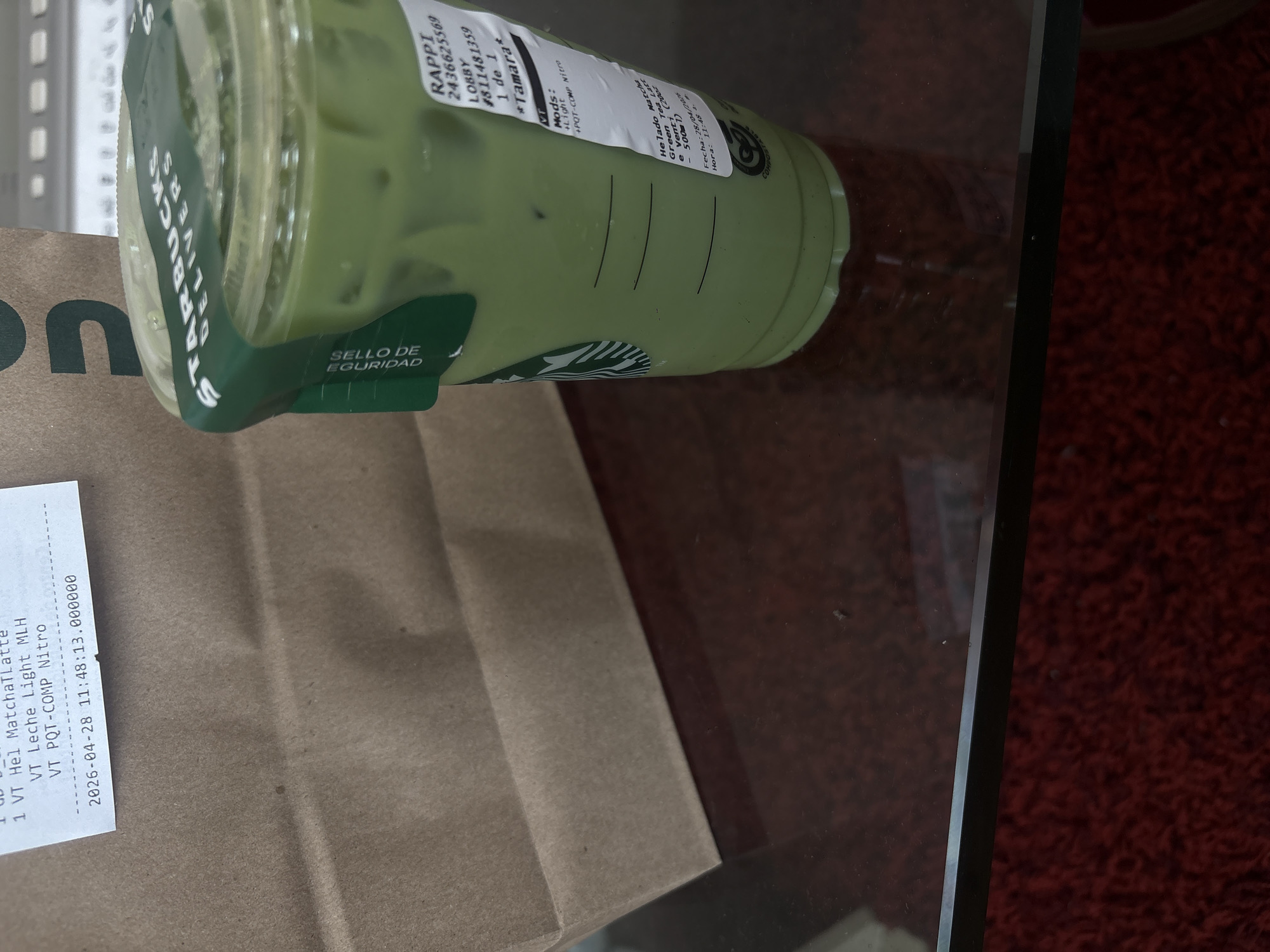 This close-up, overhead shot captures a Starbucks delivery order placed on a transparent glass table in what appears to be an indoor setting in Mexico City, Mexico. The glass surface reflects a dark red, textured material, possibly a carpet or rug, beneath it.
The primary subject is a clear plastic Starbucks cup filled with a light green, iced matcha latte, complete with ice cubes. A dark green paper sleeve around the cup features the "STARBUCKS DELIVERS" logo and a "SELLO DE SEGURIDAD" (security seal) tab. Attached to the cup is a white sticker with "RAPPI" branding, indicating it was delivered by the Rappi service. The sticker also shows an order number "2436625569", "LOBBY", a reference number "#811481359", and the recipient's name "*Tamara*". Drink details include "Helado Matcha," "Green Tea Latt e Venti (2002 500ml)", and modifications: "+Light" and "+POT-COMP Nitro". The cup also bears a "COMPOSTABLE" label.
Beside the cup, a brown paper bag is partially visible, with a fragment of a letter "on" visible, likely part of a delivery service logo. A white receipt, possibly from Starbucks or the delivery platform, is taped to the bag. It lists "1 VT Hel MatchaLatte", "VT Leche Light MLH", and "VT PQT-COMP Nitro", corresponding to the drink and its modifications. Both the cup's label and the receipt display the date "2026-04-28" and time "11:48", suggesting a future date for the order.