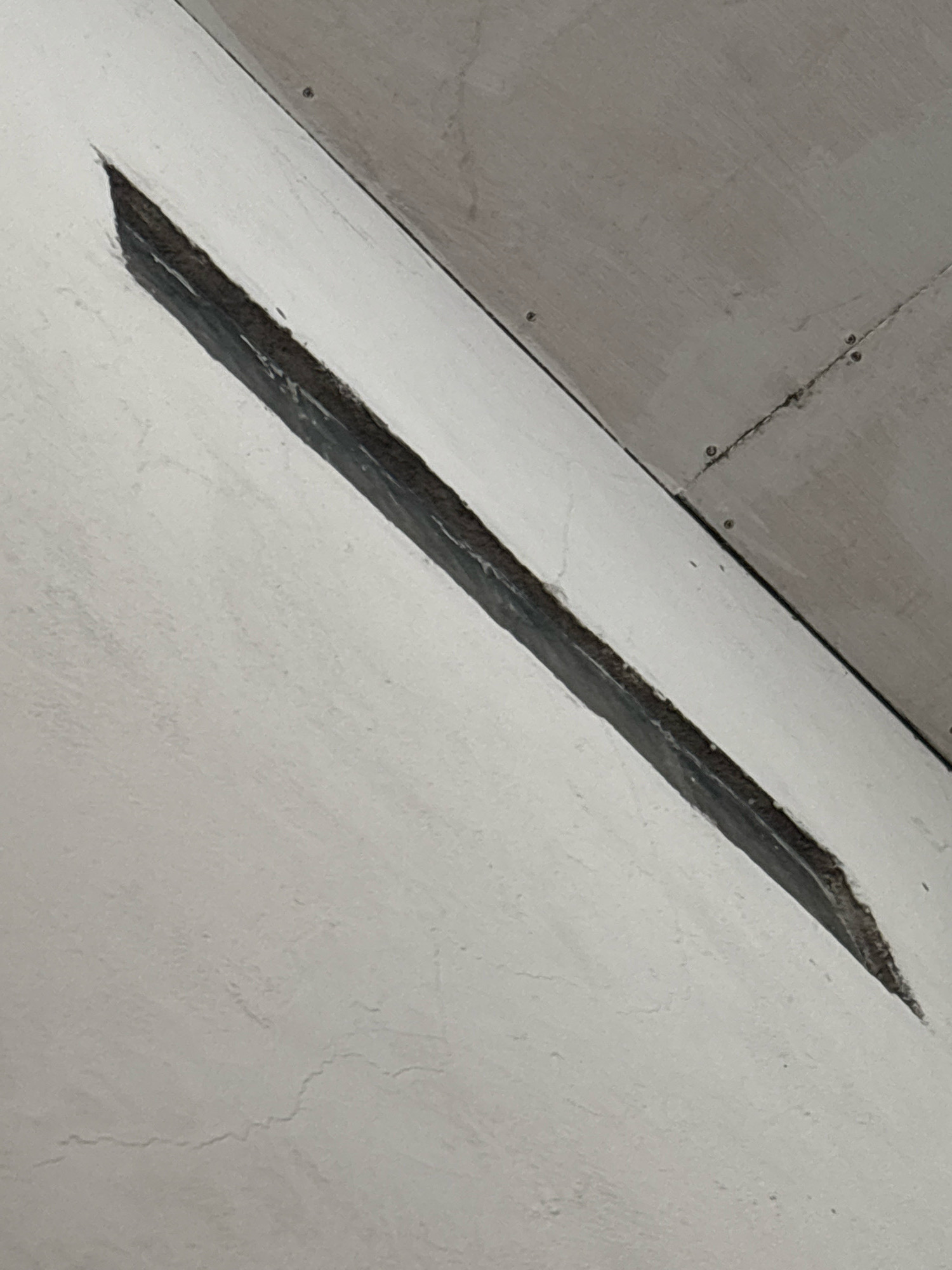 The image shows a close-up of a crack in a white painted wall, with a darker, recessed area within the crack. The wall appears to be part of an interior space, possibly a ceiling or an angled wall. The texture of the wall is uneven, with some visible imperfections. Above the white wall, a grey surface is visible, which appears to be a joint or a different material, also showing some texture and screw heads. The lighting in the image is somewhat dim, casting a shadow within the crack. There are no people or objects depicted in the image. This image is not from North Jakarta, Indonesia.