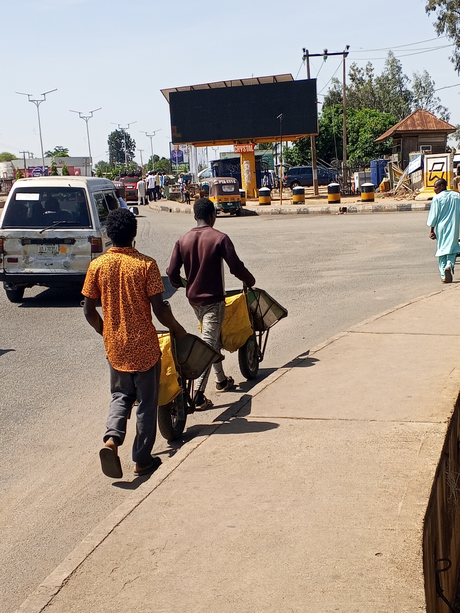 This outdoor image captures a sunny street scene in an urban or semi-urban environment. The sky is clear, and strong shadows suggest it is midday or early afternoon.
In the foreground, two men walk along a paved sidewalk, each pushing or pulling a modified wheelbarrow or pushcart. The man closest to the viewer wears an orange and black patterned short-sleeved shirt, dark trousers, and flip-flops. Ahead of him, the second man wears a dark long-sleeved shirt, light-colored trousers, and sneakers. Both carts are covered with yellow fabric or tarpaulin, indicating they are likely transporting goods.
Further ahead on the right side of the road, a third man walks away from the camera, dressed in a light blue traditional garment and flip-flops.
To the left, a white van or minibus is visible on the road. The background reveals a busy intersection or roundabout with other vehicles, including a yellow tricycle, and pedestrians. A large black billboard with a yellow support structure stands prominently in the mid-ground, with the word "CRYSTAL" clearly visible on its base. Numerous modern streetlights are dispersed throughout the background. On the far right, a small building with a reddish-brown corrugated metal roof is nestled among green trees. The scene depicts a lively street with ongoing daily activities.
