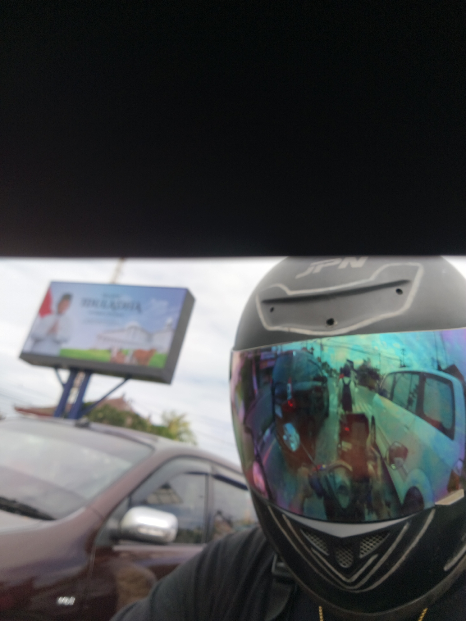 The image captures a close-up of a person wearing a black motorcycle helmet with a reflective visor. The visor reflects a busy street scene with various vehicles, including cars and motorcycles, caught in traffic. The helmet has the "JPN" logo visible on its top. In the background, a billboard with a political advertisement is partially visible, featuring a person in a white shirt and a red and white flag. To the left, the hood and side of a maroon car are in the foreground, suggesting the perspective is from a vehicle. The sky appears overcast. The specific location in Denpasar, Indonesia is not discernible from the visual elements alone.