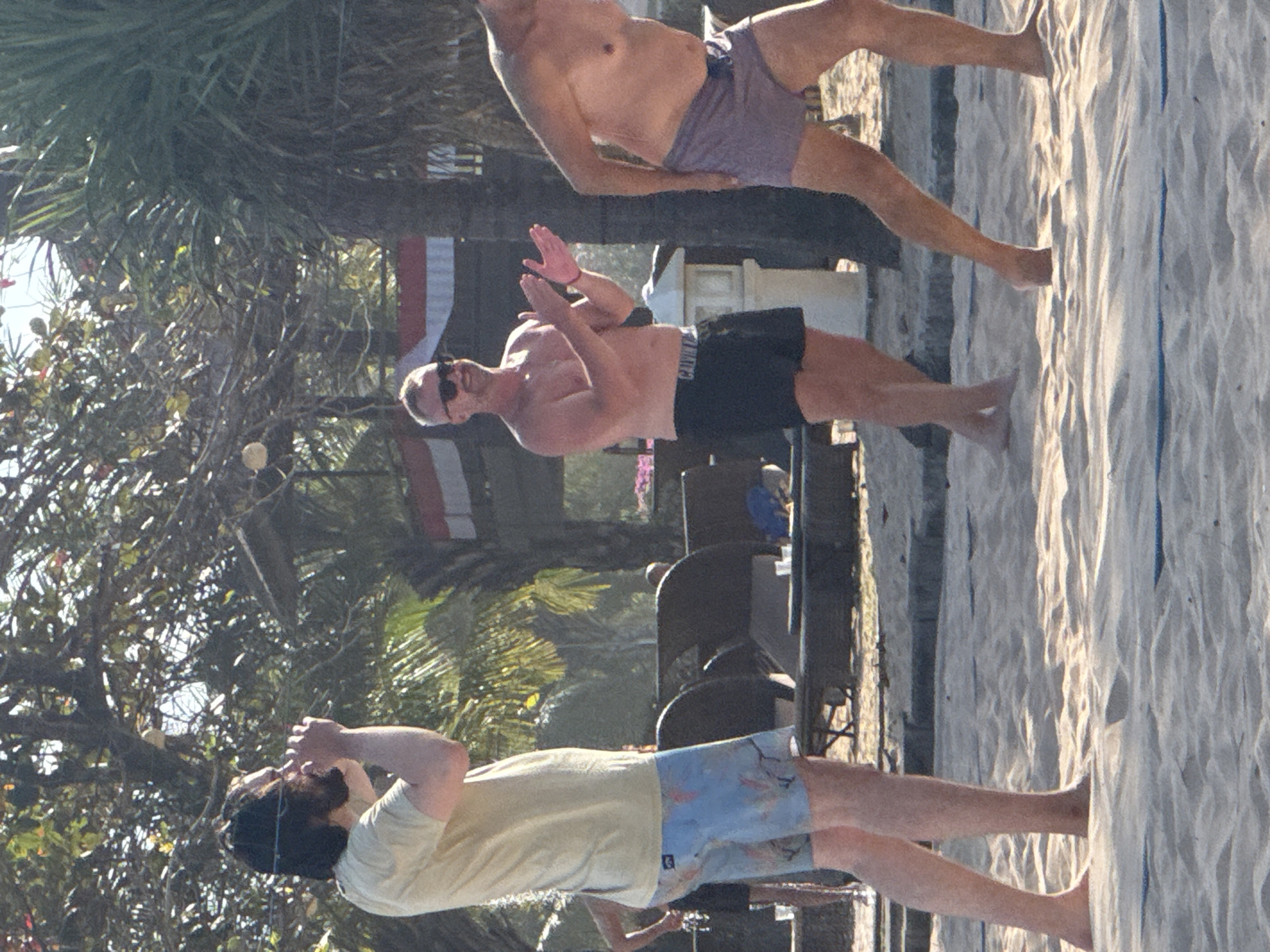The image depicts a sunny beach scene in Kuta Selatan, Indonesia, with three men visible. In the foreground and to the left, a man wearing a light yellow t-shirt and patterned blue shorts stands facing left, with his hands cupped to his mouth as if signaling or shouting. Behind him and slightly to the right, another man is clapping his hands, wearing sunglasses and black swim trunks. To the far right, a third man, with his back partially to the camera, is wearing purple swim trunks. Palm trees and tropical foliage are present in the background, along with lounge chairs and what appears to be part of a beachside structure. The sand is textured, indicating a beach environment.