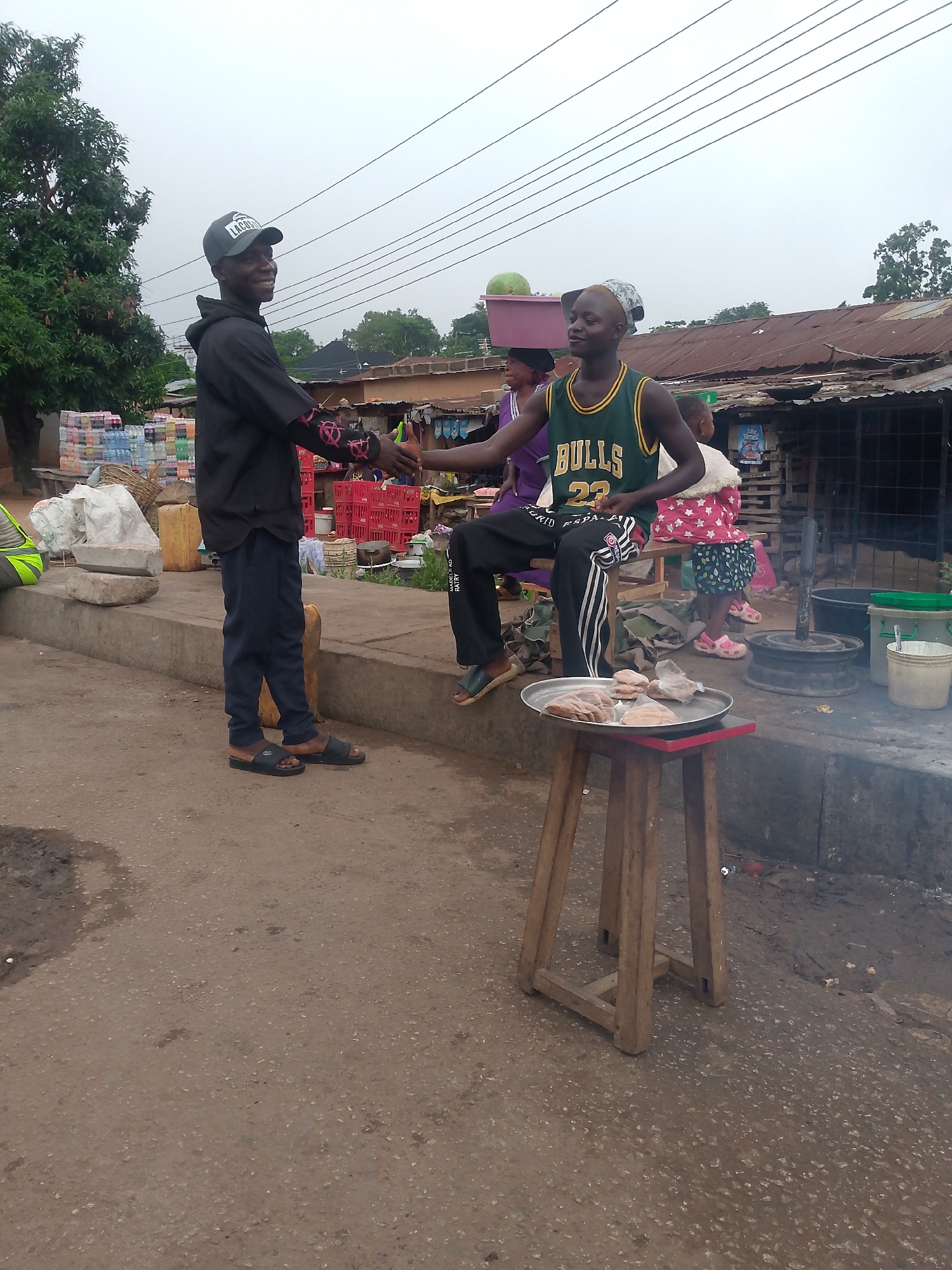 The image captures an outdoor scene in Bukuru, Nigeria. Two young men are shaking hands. One is standing, wearing a black hoodie with a "Lacoste" cap, and dark trousers. The other is seated on a stool, wearing a "Bulls 23" basketball jersey and Adidas pants. A tray of what appears to be cooked food is placed on a smaller stool next to him. In the background, there are stacks of goods, red crates, and a woman carrying a pink container on her head. A child in a pink coat and floral skirt stands near a metal cooking apparatus. Buildings with corrugated iron roofs are visible. The ground is asphalt.
