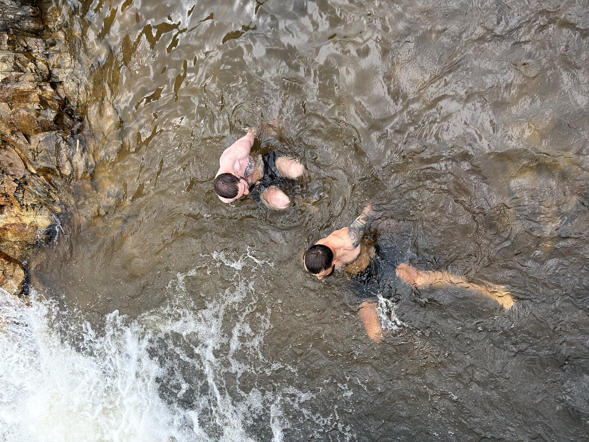 Two men are standing in shallow water near a rocky embankment and a small waterfall. The water is murky brown and has some white foam on the surface. The man in the foreground is facing away from the camera, looking towards the waterfall. He has dark hair and is wearing black swim shorts. The man in the background is also facing away from the camera, his head and shoulders visible above the water. He has short dark hair and is wearing dark swim shorts. Both men appear to be of similar build. The rocks behind them are varied in color, with shades of brown, orange, and gray. The waterfall is on the left side of the image, with white water cascading down. The overall scene suggests a natural outdoor setting, possibly a river or creek.