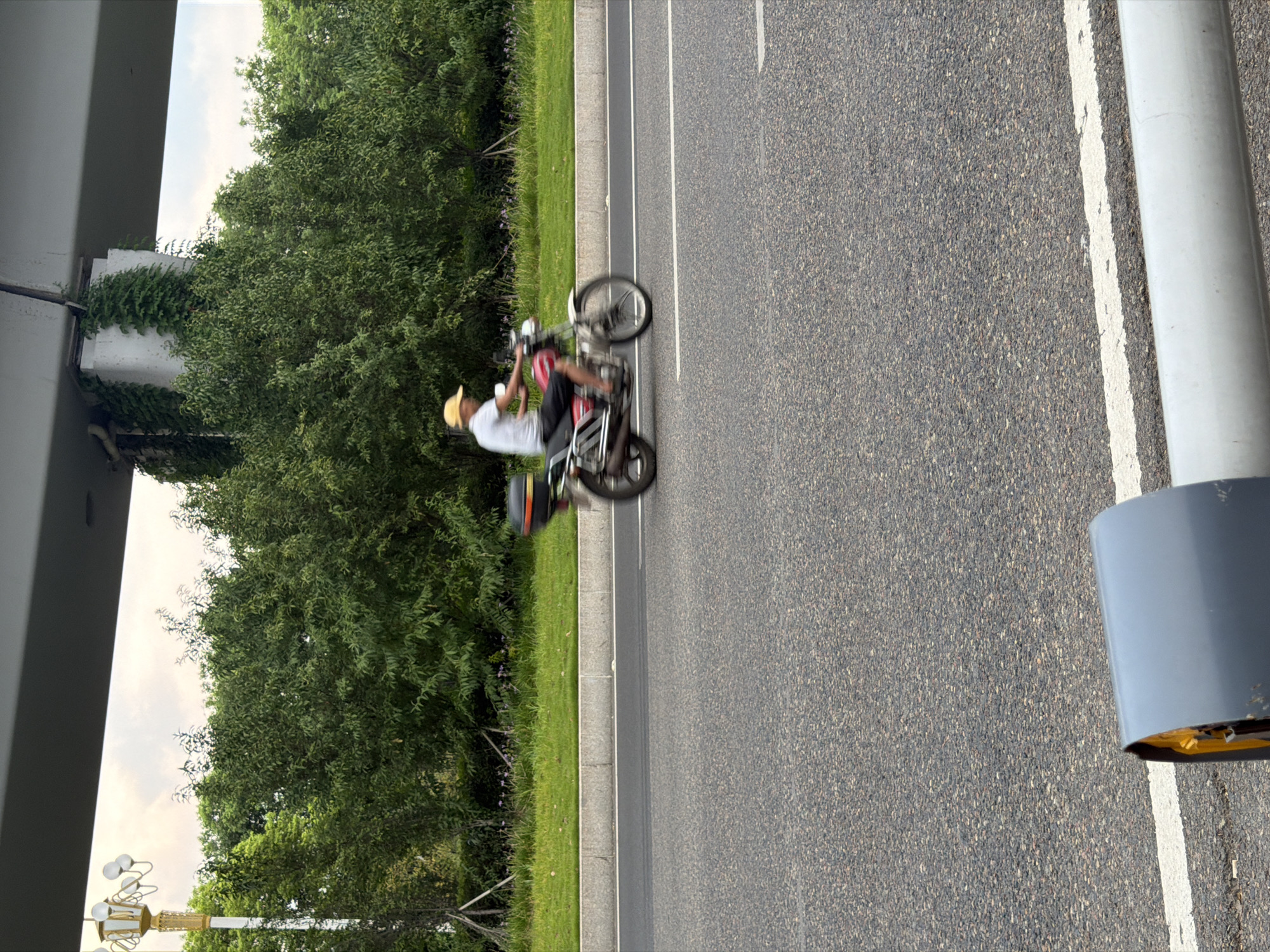 A man in a yellow hat and white t-shirt rides a red motorcycle on a paved road. He is wearing black shorts. The motorcycle has a black and red storage box on the back. The road is lined with grass and trees. An overpass is visible above the trees. The scene appears to be in Jiangjin, China.