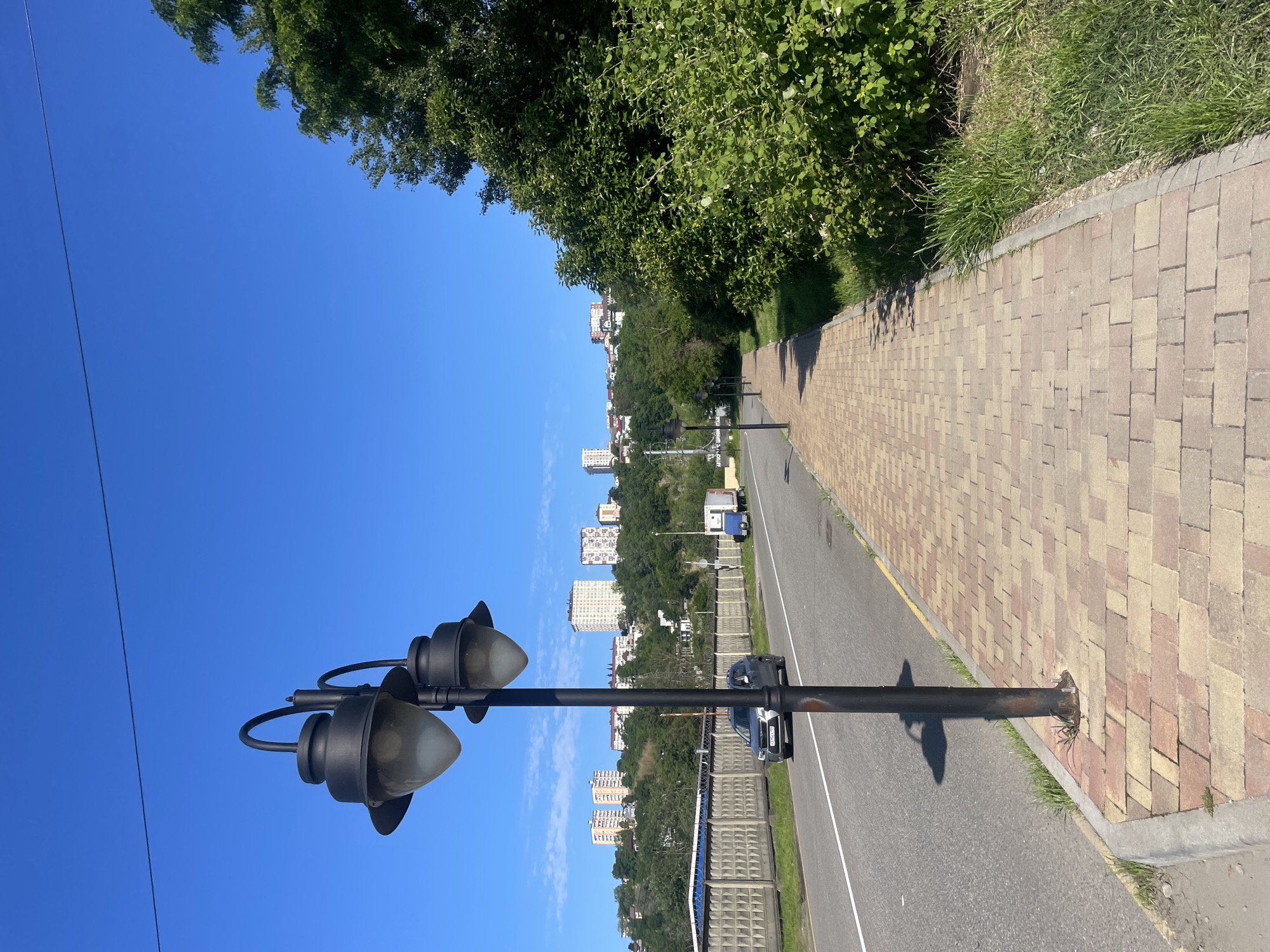 The image captures a sunny, clear day in Sochi, Russia, showcasing a blend of urban infrastructure and natural green landscapes. The scene is viewed along a paved road and sidewalk, with distant residential buildings nestled among hills.
In the foreground, a tall, black lamppost with two traditional-style lights stands prominently on the left, casting a distinct shadow onto the asphalt road. To its right, a meticulously laid sidewalk, made of alternating light and dark beige brick pavers, runs parallel to the road. Beyond the sidewalk, a narrow strip of green grass borders a thick line of lush, leafy green trees and bushes that extend into the midground.
The midground features a two-lane asphalt road, clearly marked with a solid white line on the right edge and a broken yellow line further left. A dark-colored sedan, possibly a Hyundai, is visible on the road, partially obscured by the lamppost. Its license plate, "A 555 MY 93 RUS," is discernible, with "93 RUS" indicating the Krasnodar Krai region, which includes Sochi. Along the right side of the road, a low, grey concrete barrier or fence separates the roadway from a dense, tree-covered hillside. Other similar lampposts are visible further down the road, receding into the distance.
In the background, numerous modern, white high-rise residential buildings are scattered across the rolling, green hillsides. The sky above is a brilliant, unblemished blue, with only a few faint wisps of white cloud, underscoring the bright and clear weather conditions. The strong sunlight casts sharp shadows, indicating a time around morning or early afternoon.