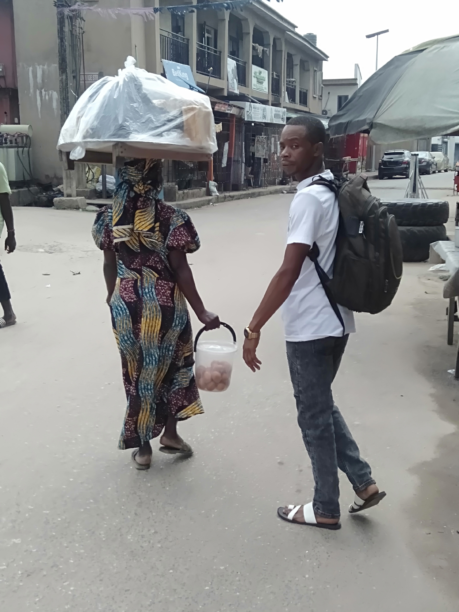 The image shows a street scene in Shangisha, Nigeria. A woman is walking away from the camera, carrying a large load wrapped in plastic on her head, balanced on a wooden frame. She is wearing a colorful patterned dress and sandals, and carrying a clear bucket filled with brown objects, likely eggs. A man is walking in the same direction as the woman, to her right. He is looking at the camera, wearing a white t-shirt, dark jeans, and sandals, and carrying a backpack. The street is paved and appears to be wet. Buildings with shops are visible in the background, along with parked cars and an umbrella. The sky is overcast.
