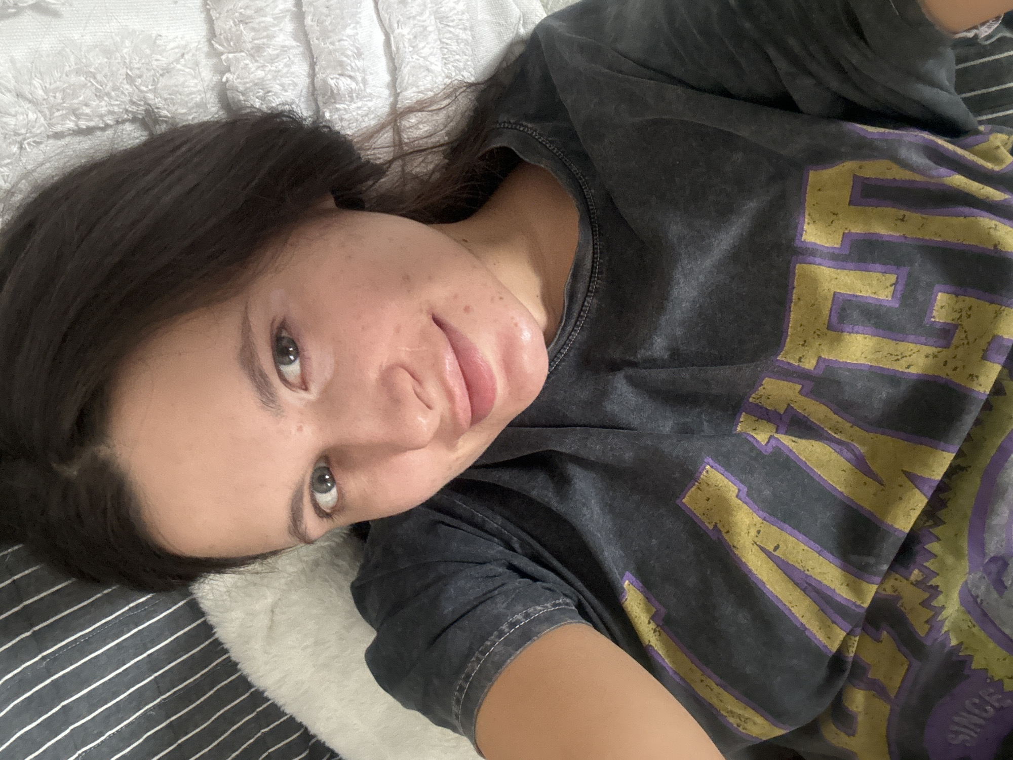 A woman with dark hair and light eyes is lying on a bed. She is wearing a grey t-shirt with gold and purple lettering. The bed has striped sheets and a white, fluffy pillow. The location is unknown and cannot be determined from the image.