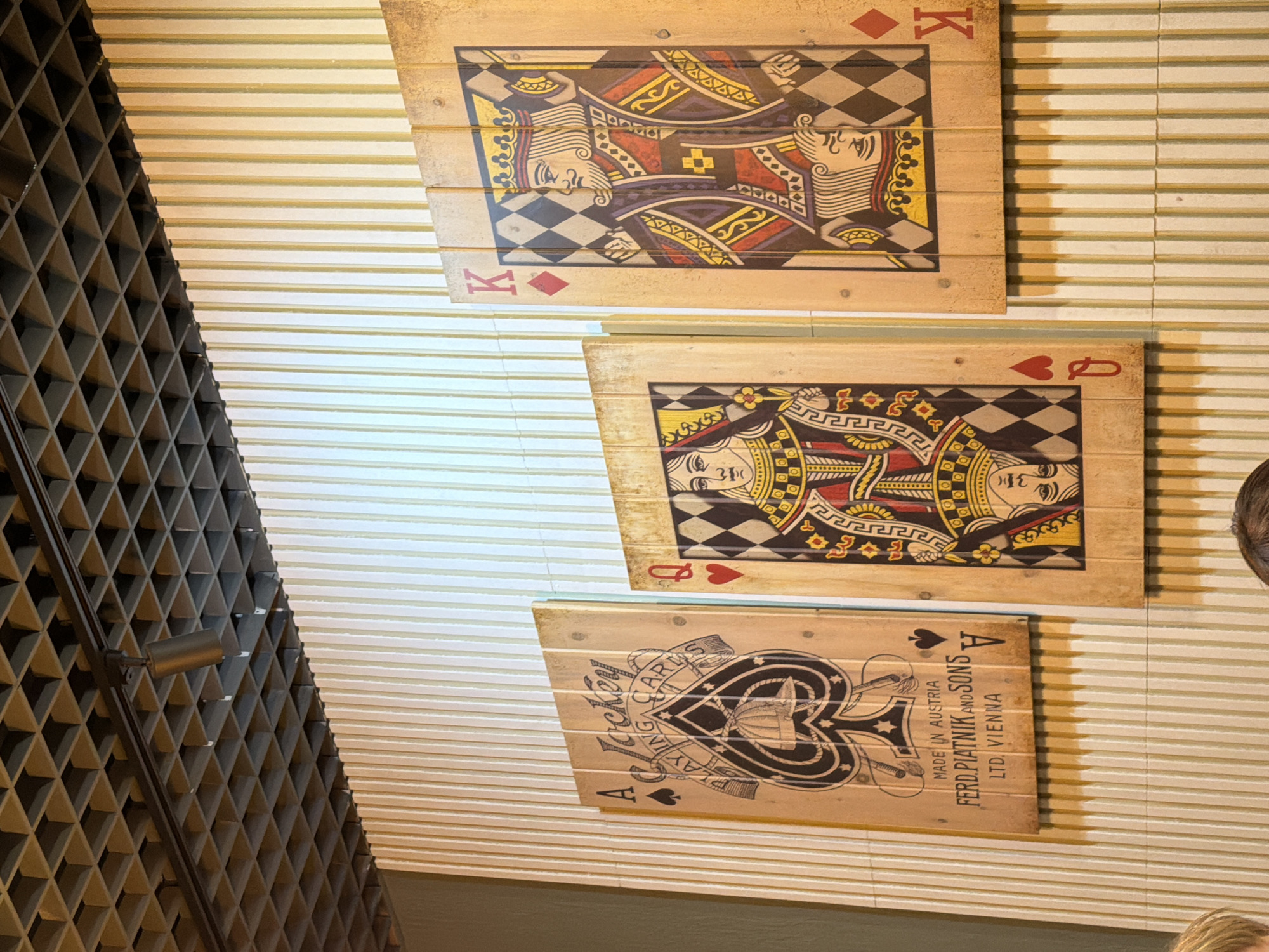 This image, captured in Chaikovsky, Russia, showcases an interior decorative display featuring three large, wooden plaques designed as playing cards. The plaques are mounted on a light-colored wall that features horizontal linear textures, resembling window blinds or paneling.
The plaques, made from joined wooden planks, have a rustic appearance. From top to bottom, they depict the King of Diamonds, the Queen of Hearts, and the Ace of Spades. The King and Queen cards feature colorful, stylized illustrations common in playing card designs, with the King wearing a crown and holding a sword, and the Queen also crowned. The Ace of Spades is depicted with a more elaborate, black and white design incorporating text and a decorative ribbon.
To the left of the plaques, a dark, intricate gridded structure, possibly a wine rack or decorative lattice, forms a sharp diagonal line across the frame. On the far right and bottom, a partial view of a person's head and hair is visible, suggesting human presence in the space.
Visible text on the plaques includes the card ranks: "K" for the King, "Q" for the Queen, and "A" for the Ace. The Ace of Spades plaque also bears detailed information: "MADE IN AUSTRIA", "FERD.PIATNIK AND SONS", "LTD. VIENNA", and "A Jockey PLAYING CARDS". The image is taken from an upward, tilted angle, giving the overall scene a dynamic perspective.