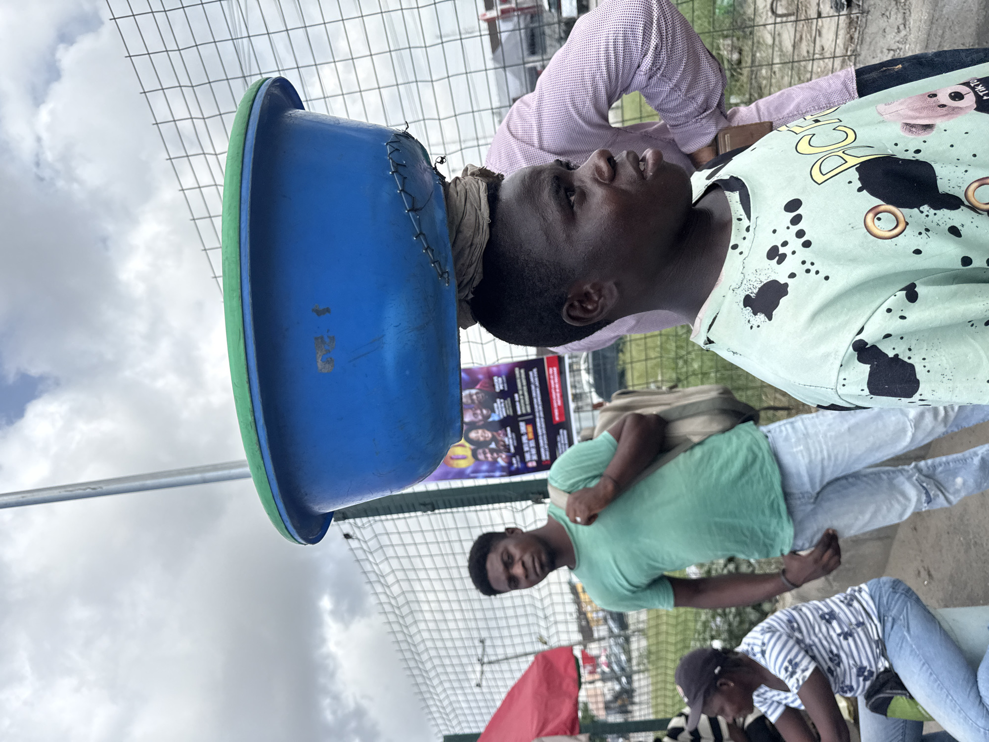 The image captures an outdoor scene in what appears to be an urban or semi-urban setting, likely in Lekki, Nigeria, during daytime under an overcast sky.
In the foreground, a young Black man is depicted, balancing a large blue plastic basin with a green rim on his head, supported by a light brown cloth. He wears a light green t-shirt adorned with black splatter patterns, yellow text, and graphics including a teddy bear and the words "TIK TOK" visible on his shoulder. His expression is neutral, and he gazes slightly upwards and to his right.
Behind him and slightly to his left, another young Black man stands, wearing a plain light green t-shirt and light blue jeans, with a beige backpack over one shoulder. He looks directly towards the viewer. Partially visible in the lower-left corner is a third individual, possibly female, wearing a striped blue and white shirt, light blue jeans, and a dark cap, appearing to be bending down. Another person in a pink checkered shirt is partly seen behind the man carrying the basin.
A wire mesh fence runs across the mid-ground, separating the subjects from the background elements. Through the fence, a large billboard or poster displaying images of people and unreadable text, a silver metal pole, a red umbrella, and indistinct vehicles and structures are visible. The scene suggests everyday activity, possibly a market or street environment, where goods are transported on the head.