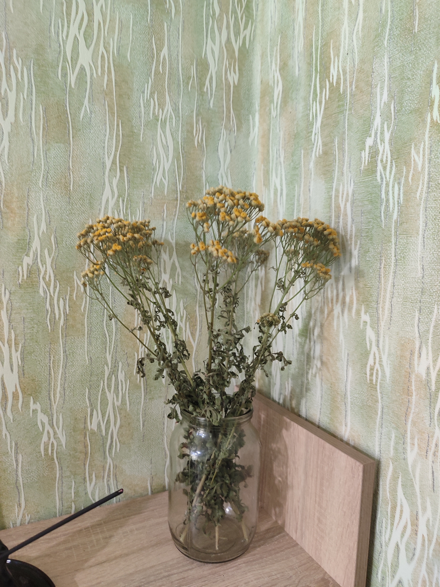 A dried bouquet of yellow wildflowers is arranged in a clear glass jar on a wooden shelf. The bouquet is positioned in front of a green and white patterned wallpaper. A narrow, dark object, possibly an incense stick, rests diagonally on the shelf to the left of the bouquet. To the right of the bouquet, a light-colored wooden panel stands vertically. The scene appears to be an indoor setting, possibly a home or a public space in the city of Lozuvatka, Ukraine. There are no people or animals visible in the image.