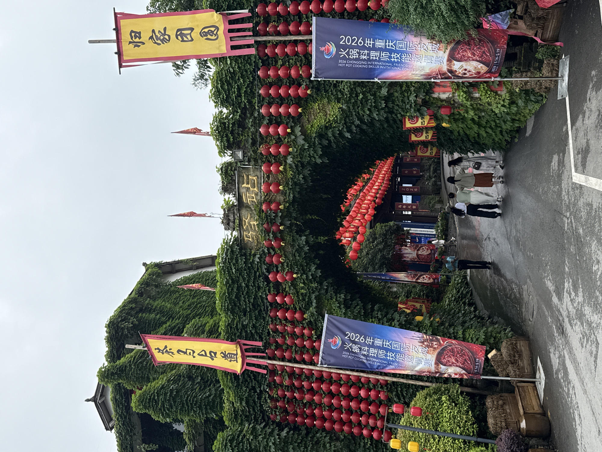 Here is a detailed description of the image:
The image shows an outdoor scene in the Nan'an District, China. Lush green foliage covers buildings and arches, creating a tunnel-like entrance. Rows of red lanterns decorate the pathway. Banners display the words "2026 Chongqing International Friendship Cities Hot Pot Cooking Skills Invitational Competition," suggesting a food-related event. People are walking along the path. Flags with Chinese characters are also present, one reading "Cha Ma Ming Dao" (Tea Horse Ancient Road). The overall atmosphere is festive and cultural.