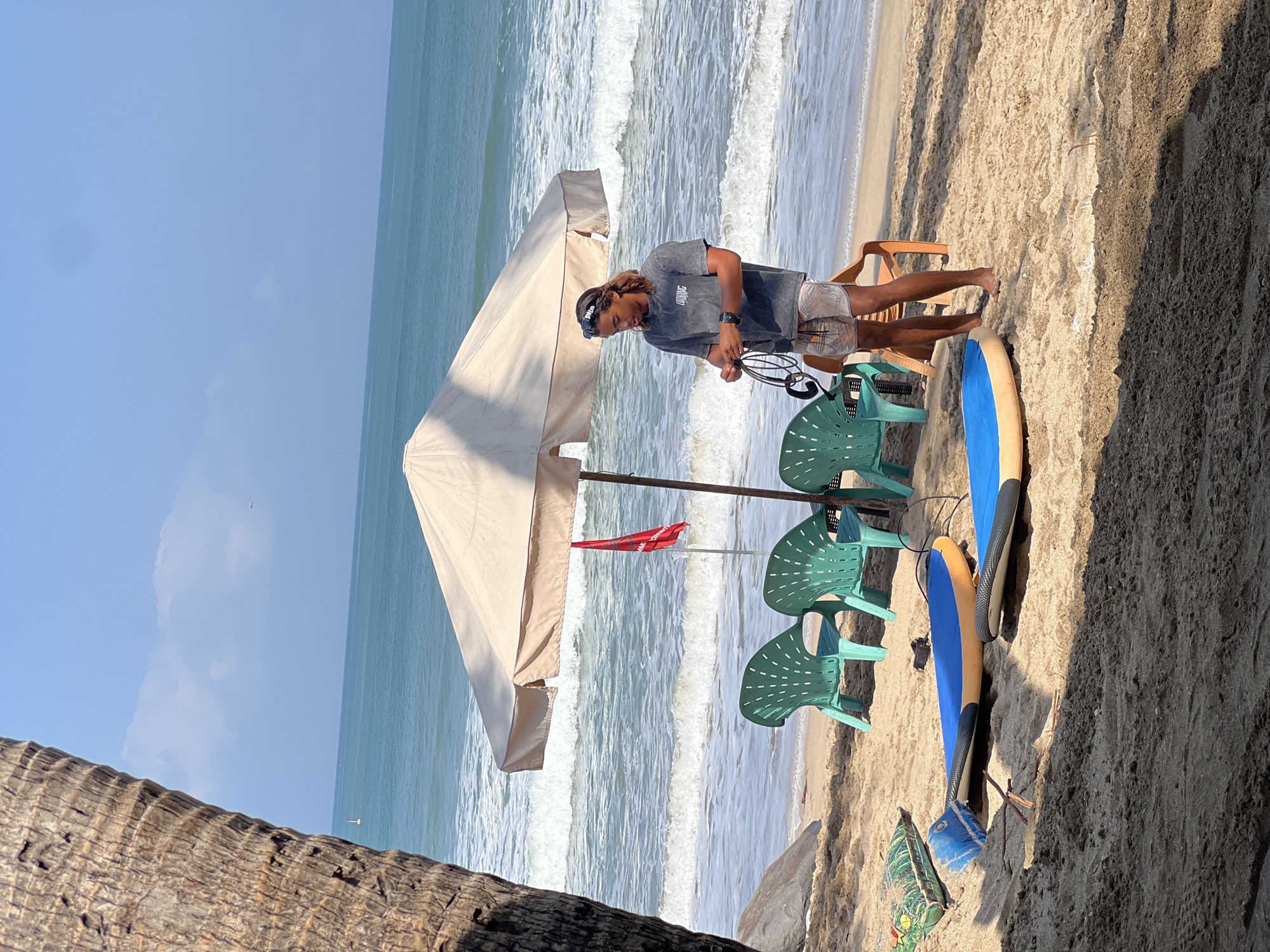 In Kuta, Indonesia, on a sandy beach, a young man with long, light brown hair and sunglasses stands under a large, cream-colored umbrella. He is wearing a grey t-shirt with a logo and patterned shorts, and is holding a coiled black leash in his hands. In front of him are two surfboards lying flat on the sand, one blue and yellow, the other blue with a black edge. To his left, three green plastic chairs are arranged, and to his right, a single orange plastic chair is visible. A small red flag with white writing, possibly a warning sign, is planted near the umbrella pole. The ocean in the background is calm with gentle waves, and the sky is clear blue with a few scattered clouds. A palm tree trunk is visible in the foreground on the left side of the image.