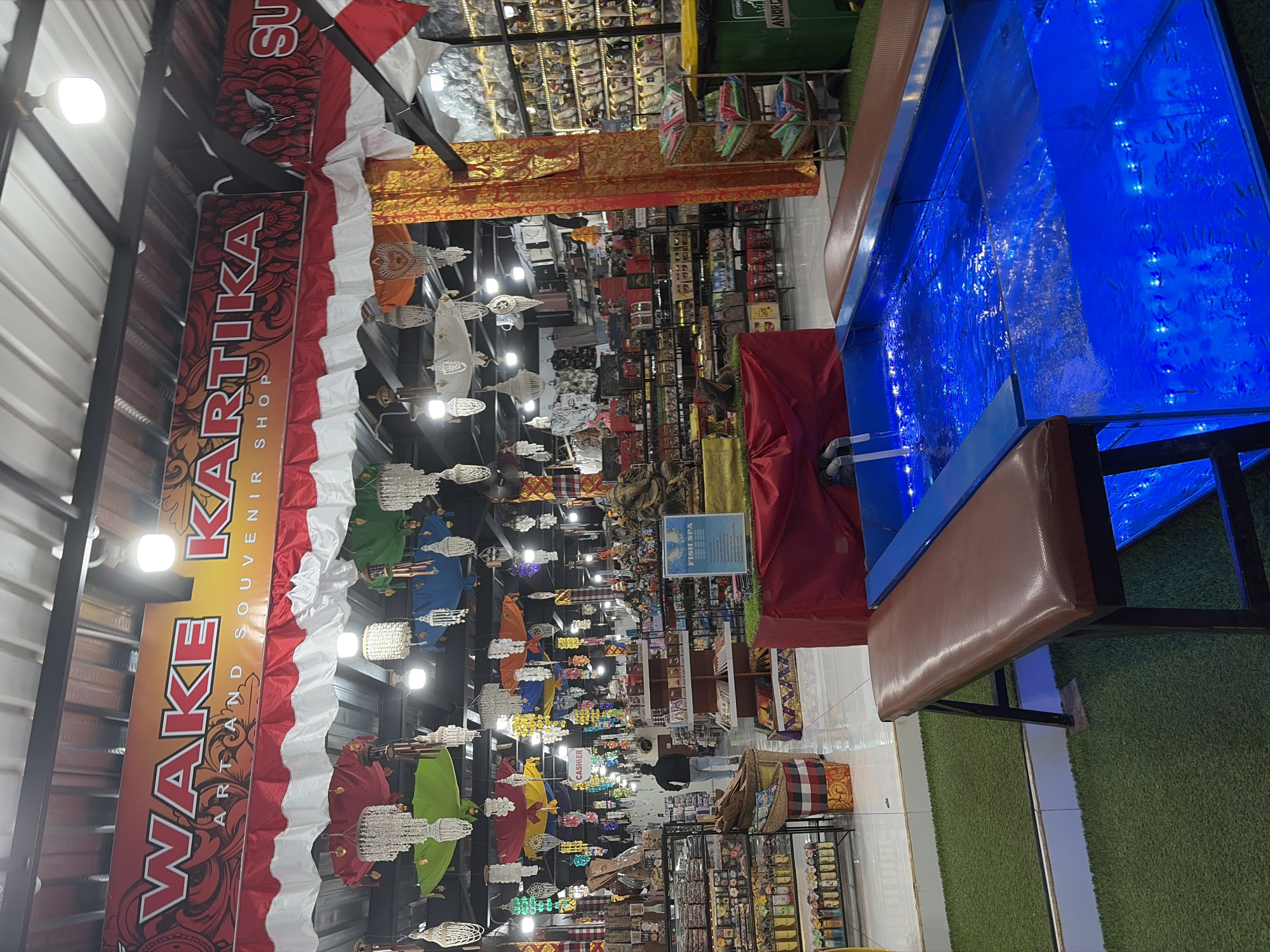 The image captures the interior of a souvenir shop, likely located in Kuta, Indonesia, as suggested by the merchandise and signage. The shop is brightly lit, with a variety of goods displayed on shelves and racks throughout. Dominating the upper portion of the image is a large banner with the text "WAKE KARTIKA ART AND SOUVENIR SHOP" in bold red lettering. Below this, colorful fabric decorations and chandeliers hang from the ceiling, adding to the festive atmosphere.
In the foreground, a clear blue fish spa tank is visible, filled with water and illuminated by blue lights, with numerous small fish swimming within. Two brown benches are positioned along the sides of the tank, suggesting it's a service offered to customers. The floor is covered with green artificial grass around the fish spa.
Further into the shop, shelves are stocked with various items, including what appear to be bottles, textiles, and packaged goods. A person is seen walking away from the camera in the middle ground, heading towards the back of the shop, where a "CASHIER" sign is visible. The environment appears to be a busy retail space, offering both souvenirs and a unique spa experience.