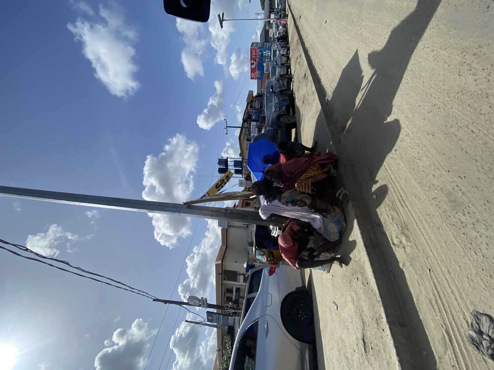A photograph shows a street scene in Ado, Nigeria. Several people are gathered under a blue umbrella next to a silver car parked along the side of the road. The sky is bright blue with white clouds. Buildings line the street and vehicles are parked or moving further down the road. Power lines stretch across the sky. The shadows of a pole and trees are cast onto the road.